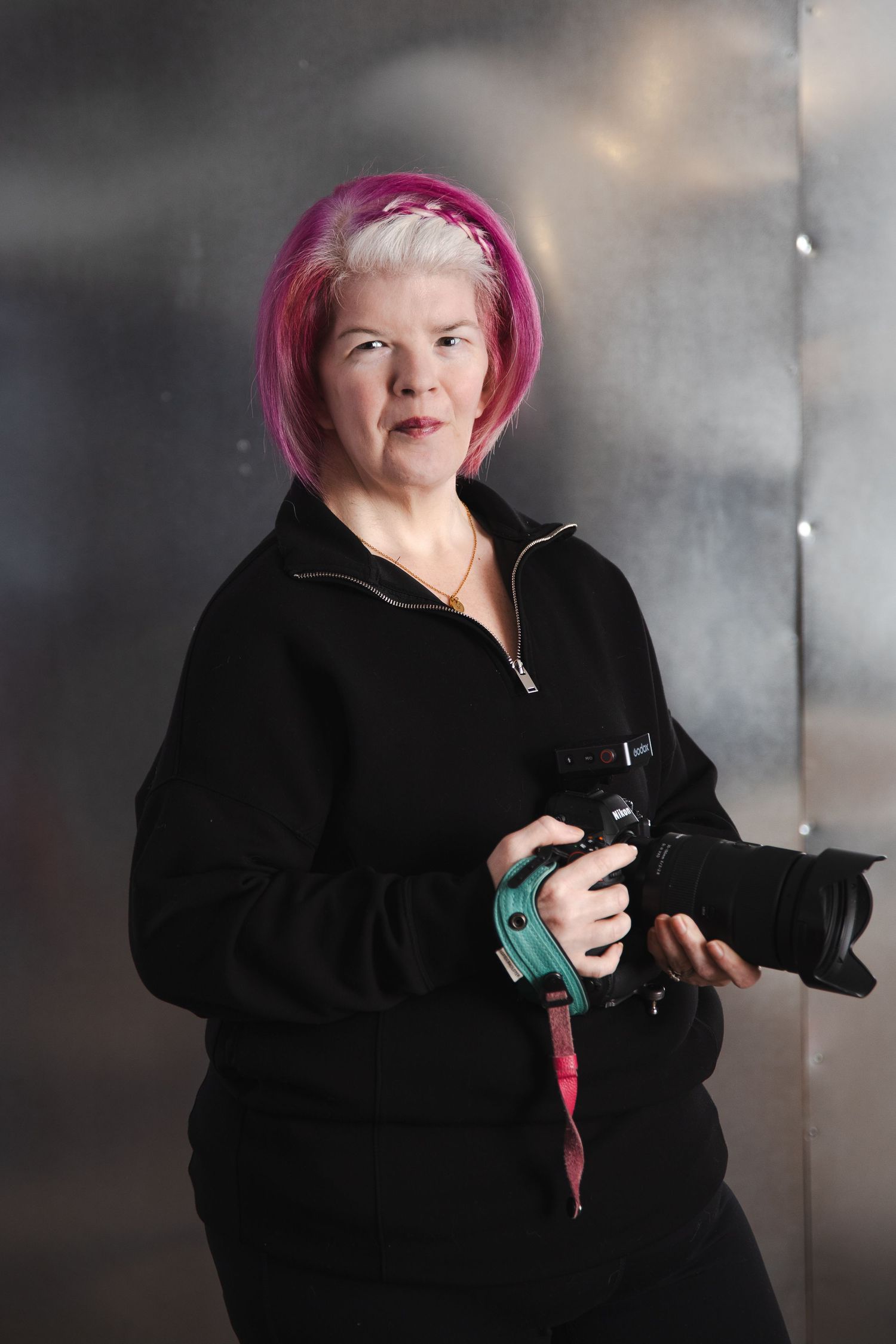Person with bright pink hair wearing a black coat poses against a gray background with a camera.
