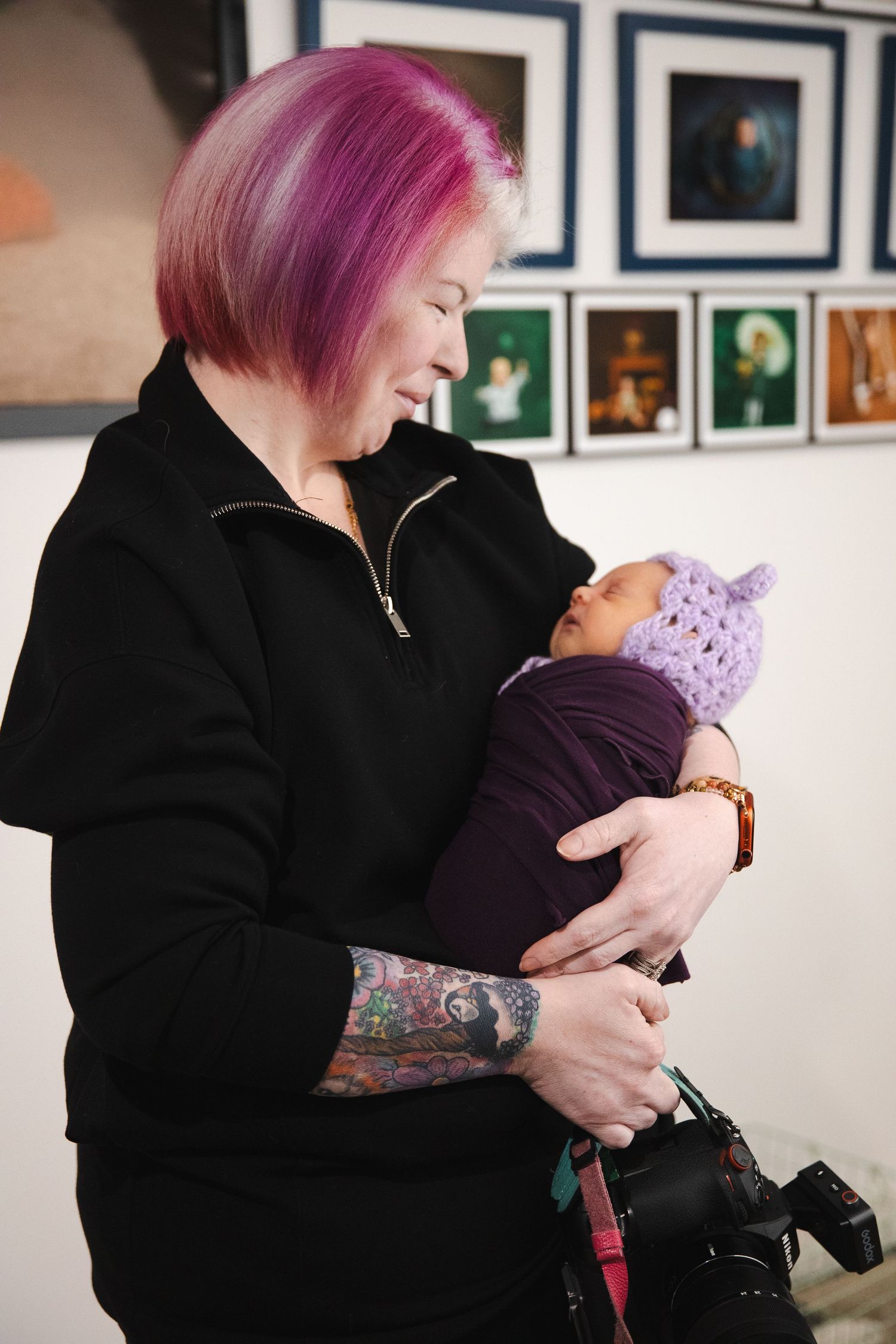 Person with pink hair in black jacket holding a small baby in a tender embrace against gallery wall background.