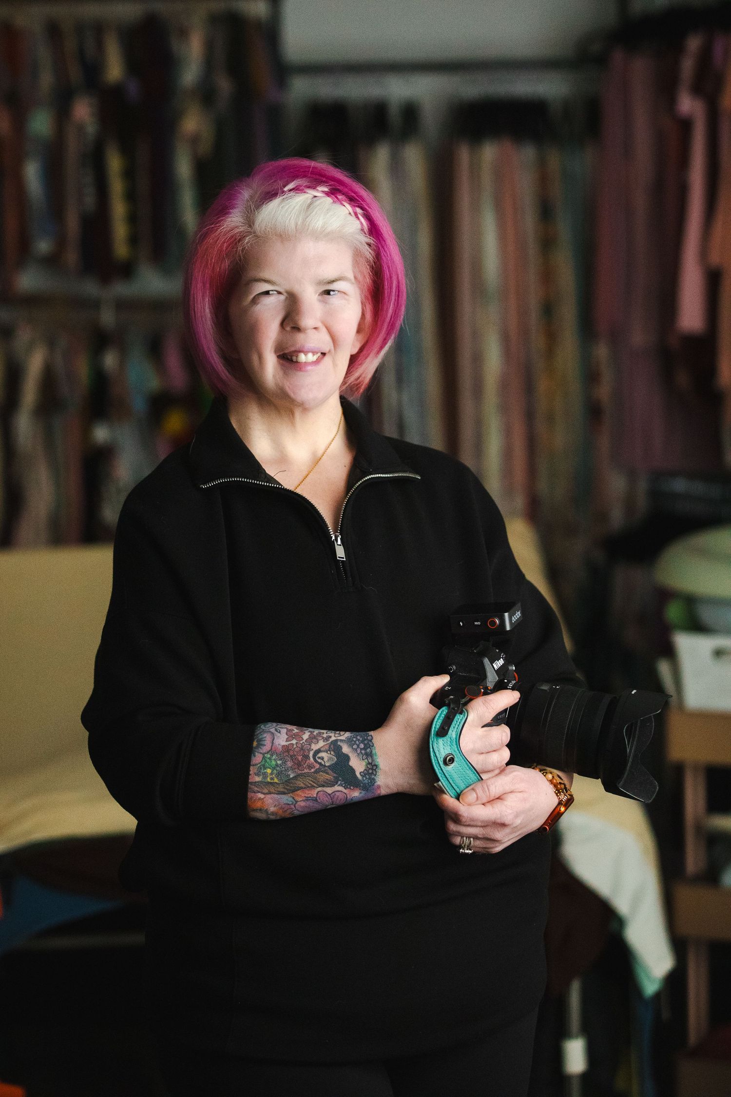 Person with bright pink hair wearing black clothing sits at desk with bookshelves in background.