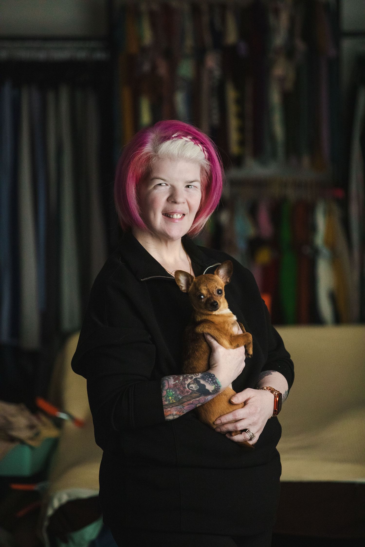 A person with pink hair wearing black holds a small brown teddy bear while smiling in a dimly lit room.