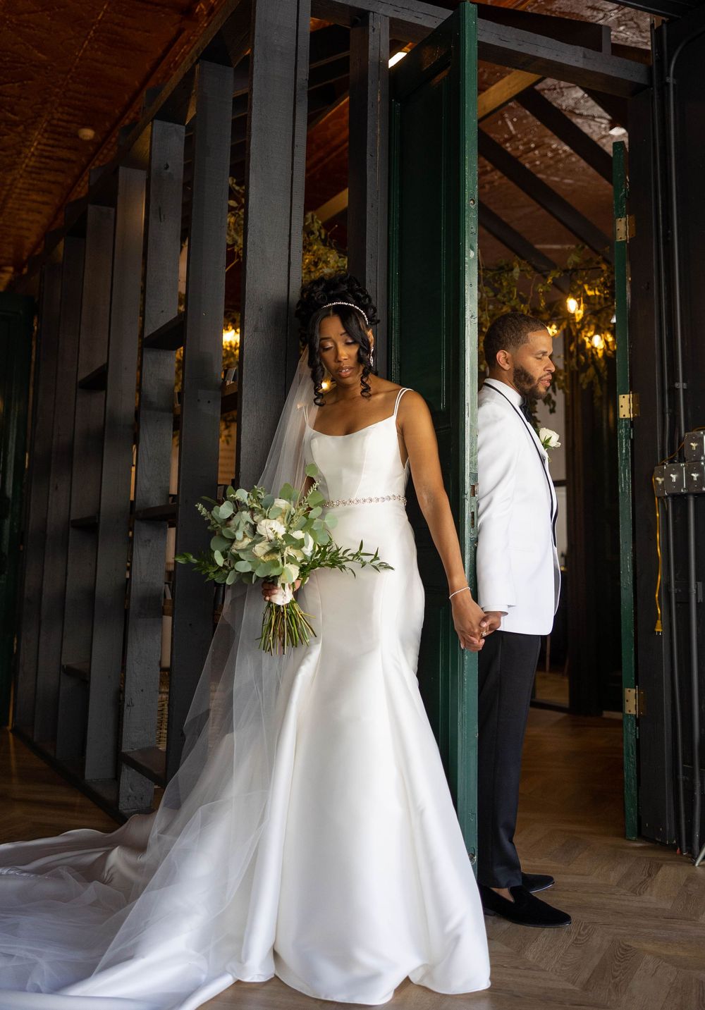 Elegant wedding couple poses in a rustic venue with wooden beams and green accents, bride holding white floral bouquet.