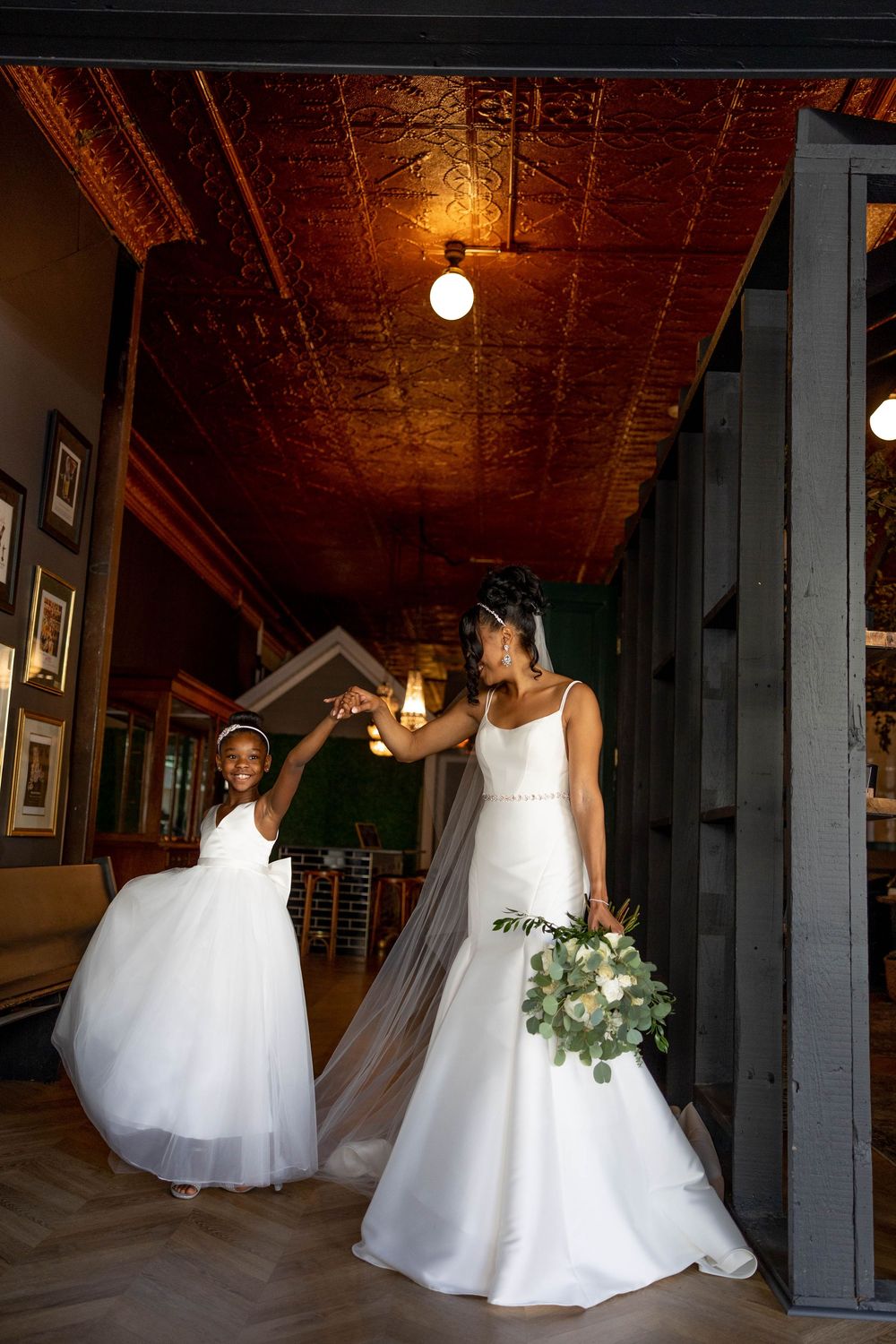 Two people in elegant white wedding dresses dance joyfully together in a rustic venue with ornate copper ceiling.