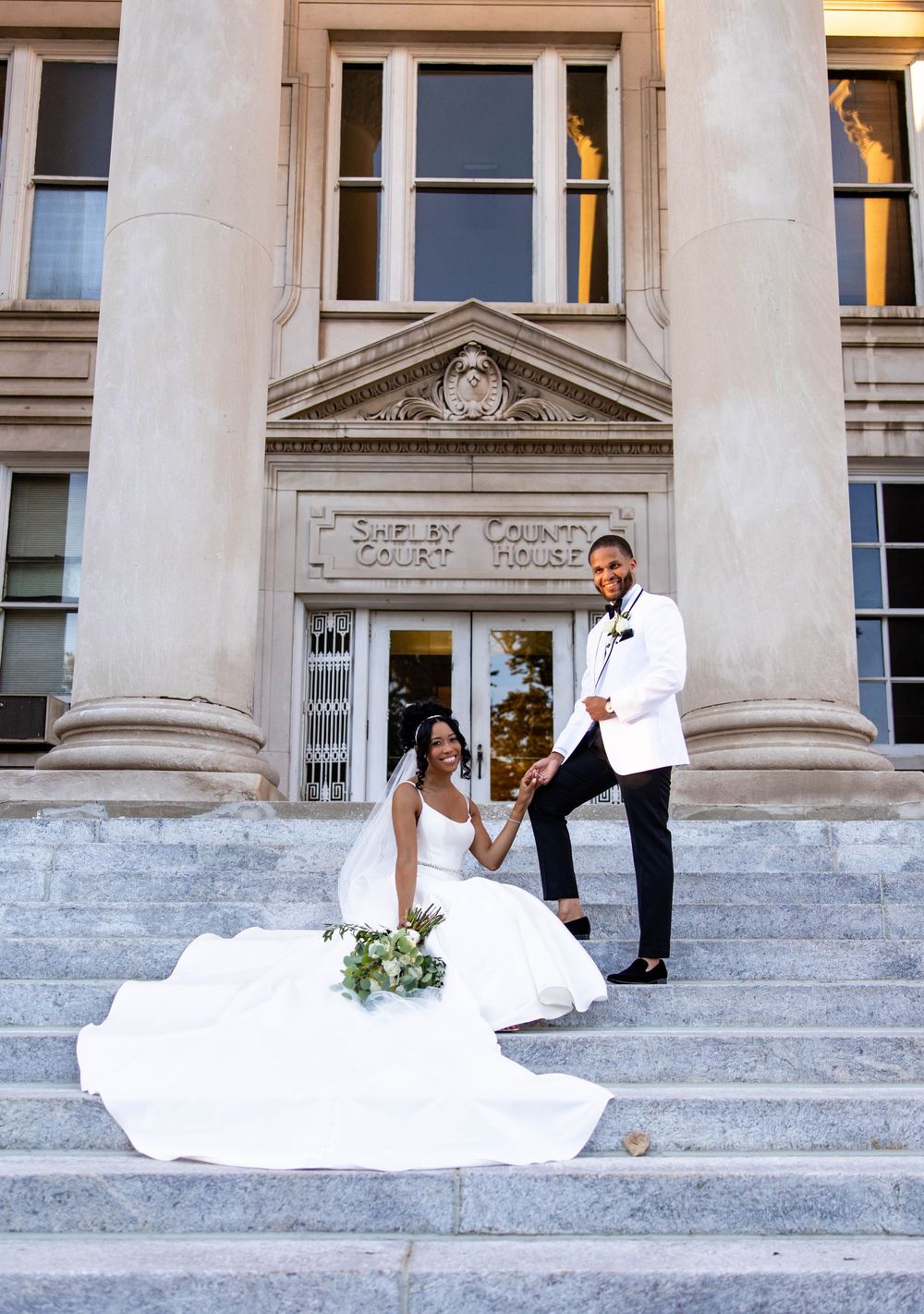 A wedding couple poses on grand stone steps of a historic building with classical columns and architectural details.