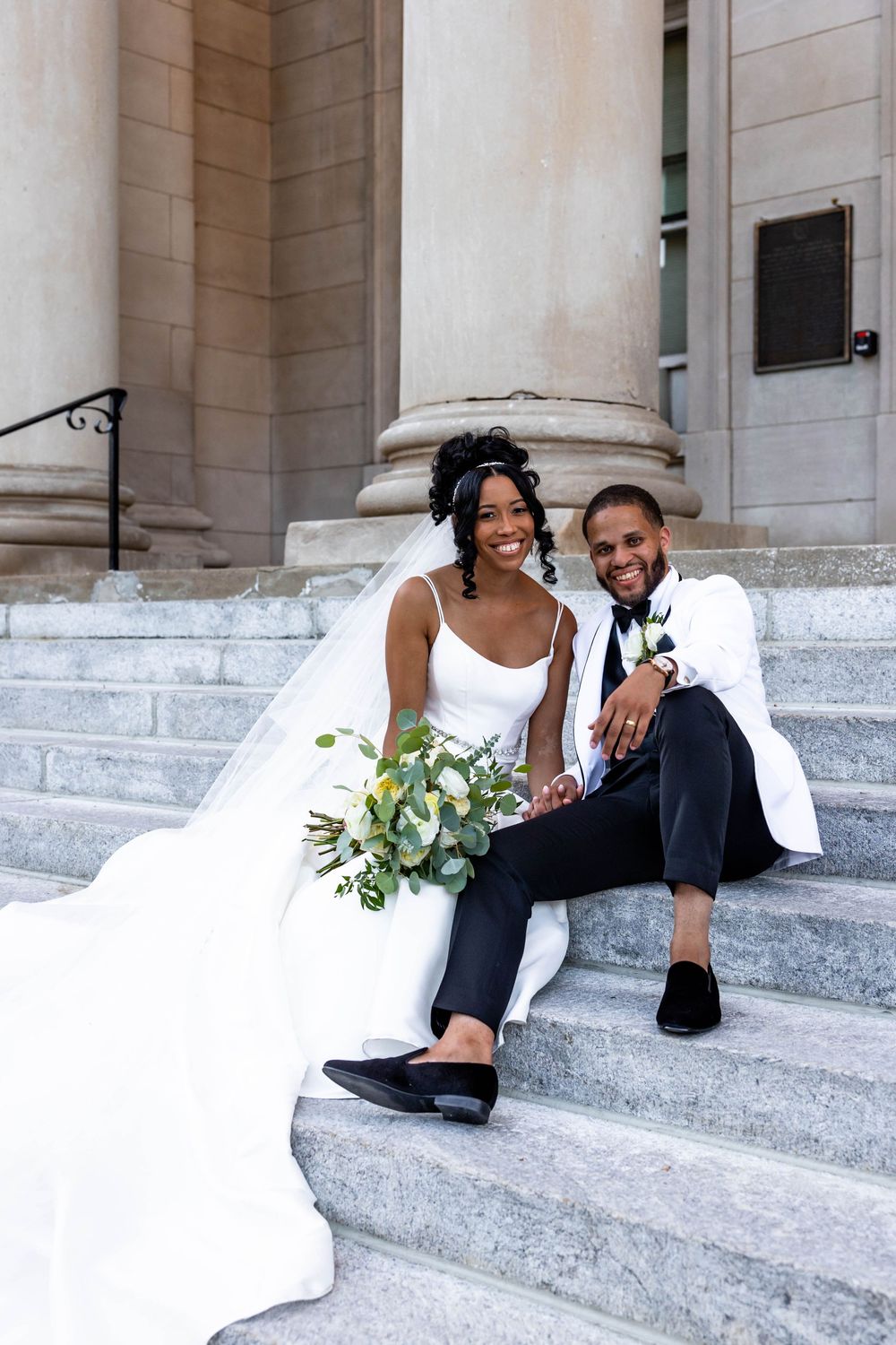 A happy couple in wedding attire poses playfully on stone steps outside a grand building.