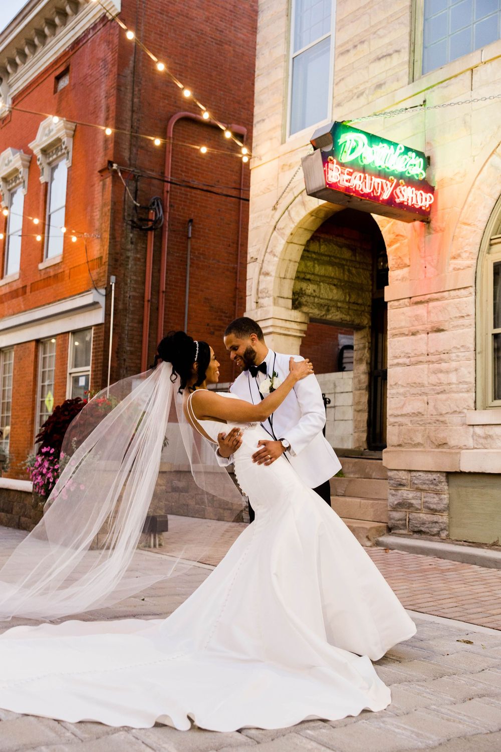 A bride in a flowing white wedding dress poses with her veil dramatically blowing in the wind against a brick building.