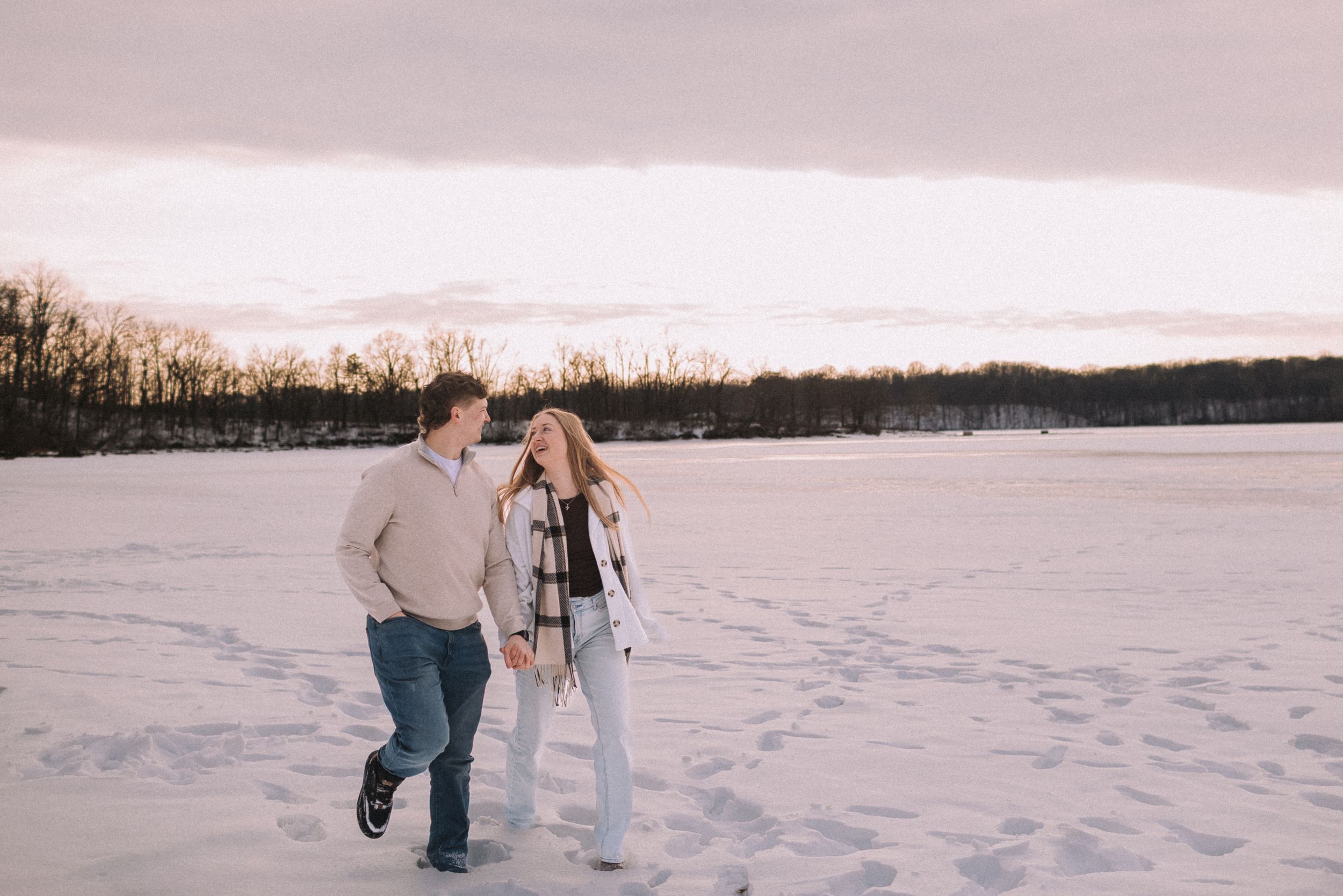 A couple walks together through a snowy winter landscape near a forest during sunset.