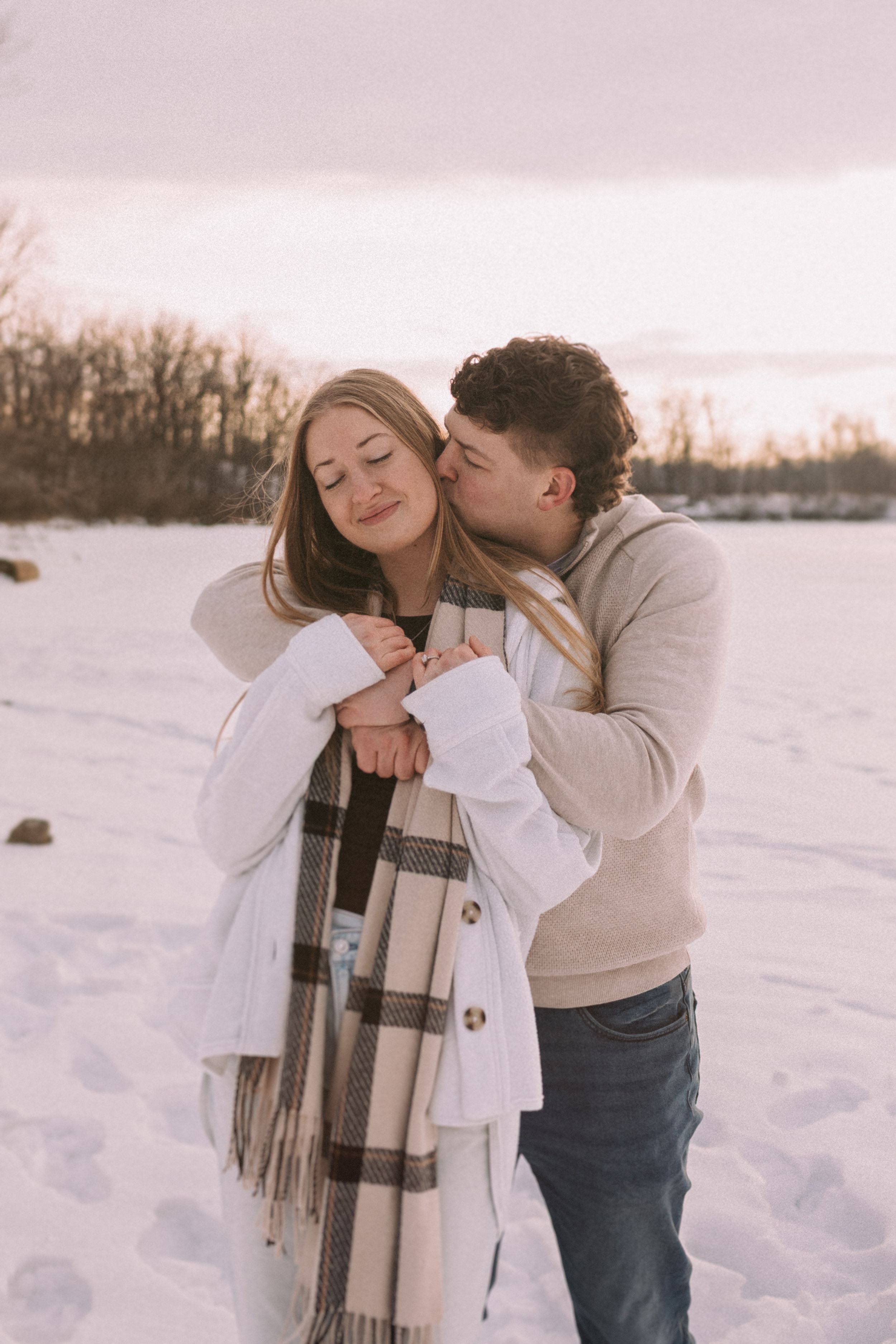 A romantic winter photoshoot featuring a couple embracing in the snow at sunset with a plaid scarf.