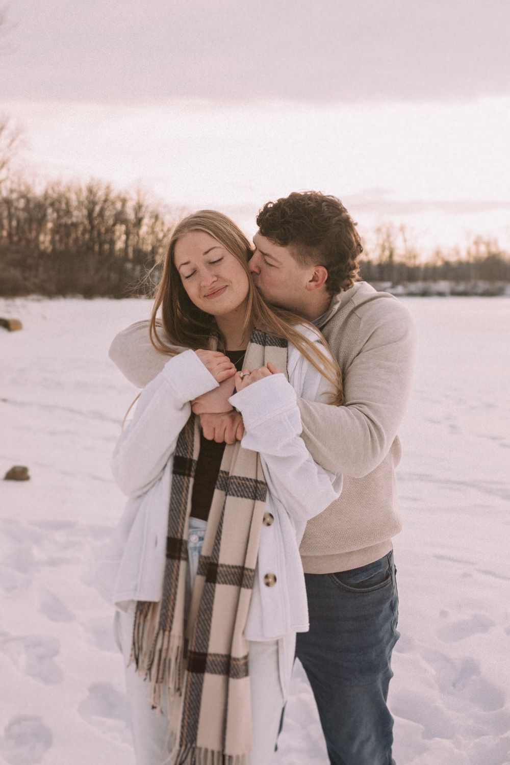A romantic winter photoshoot featuring a couple embracing in the snow at sunset with a plaid scarf.