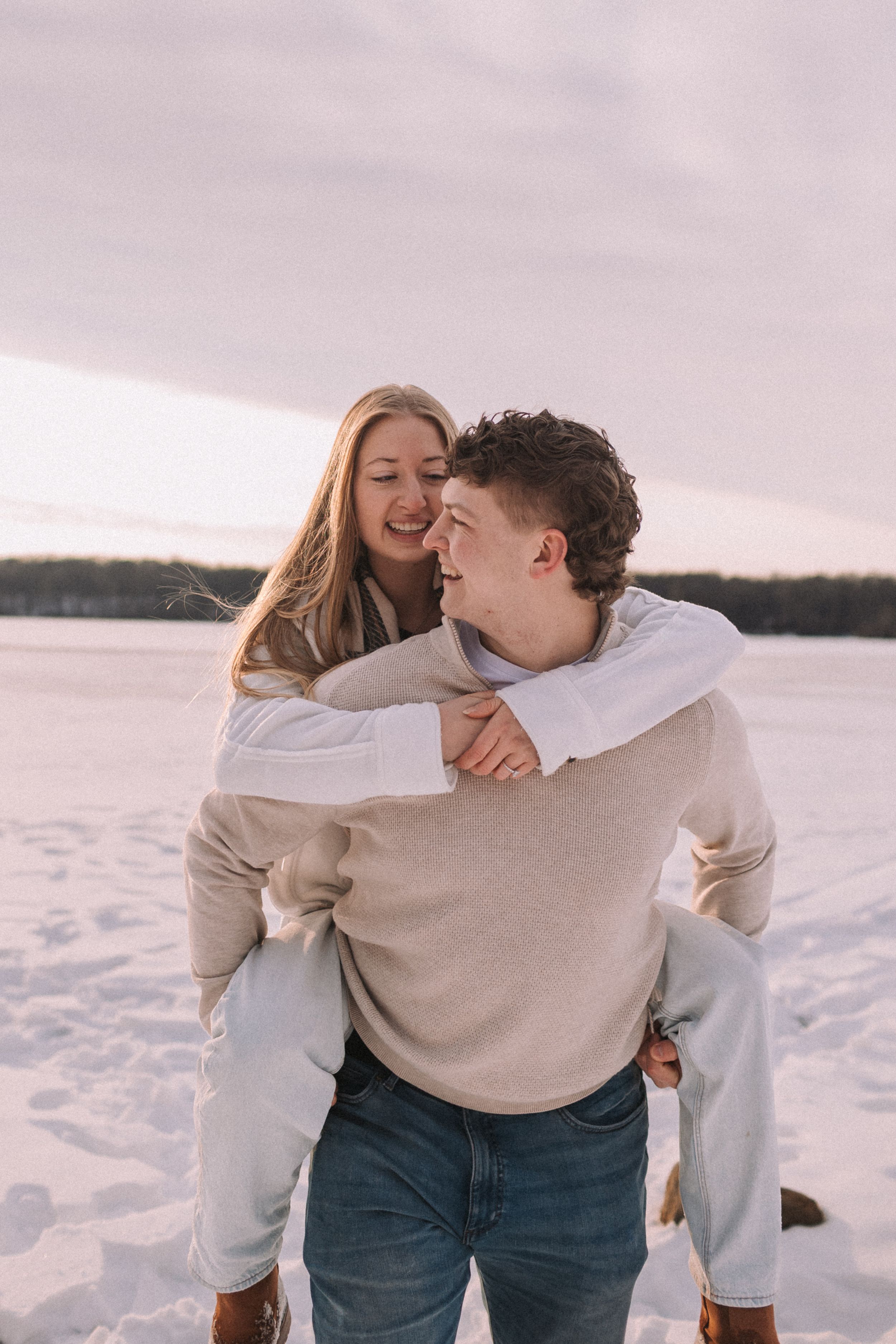A couple shares a joyful piggyback moment together in a snowy winter landscape at sunset.