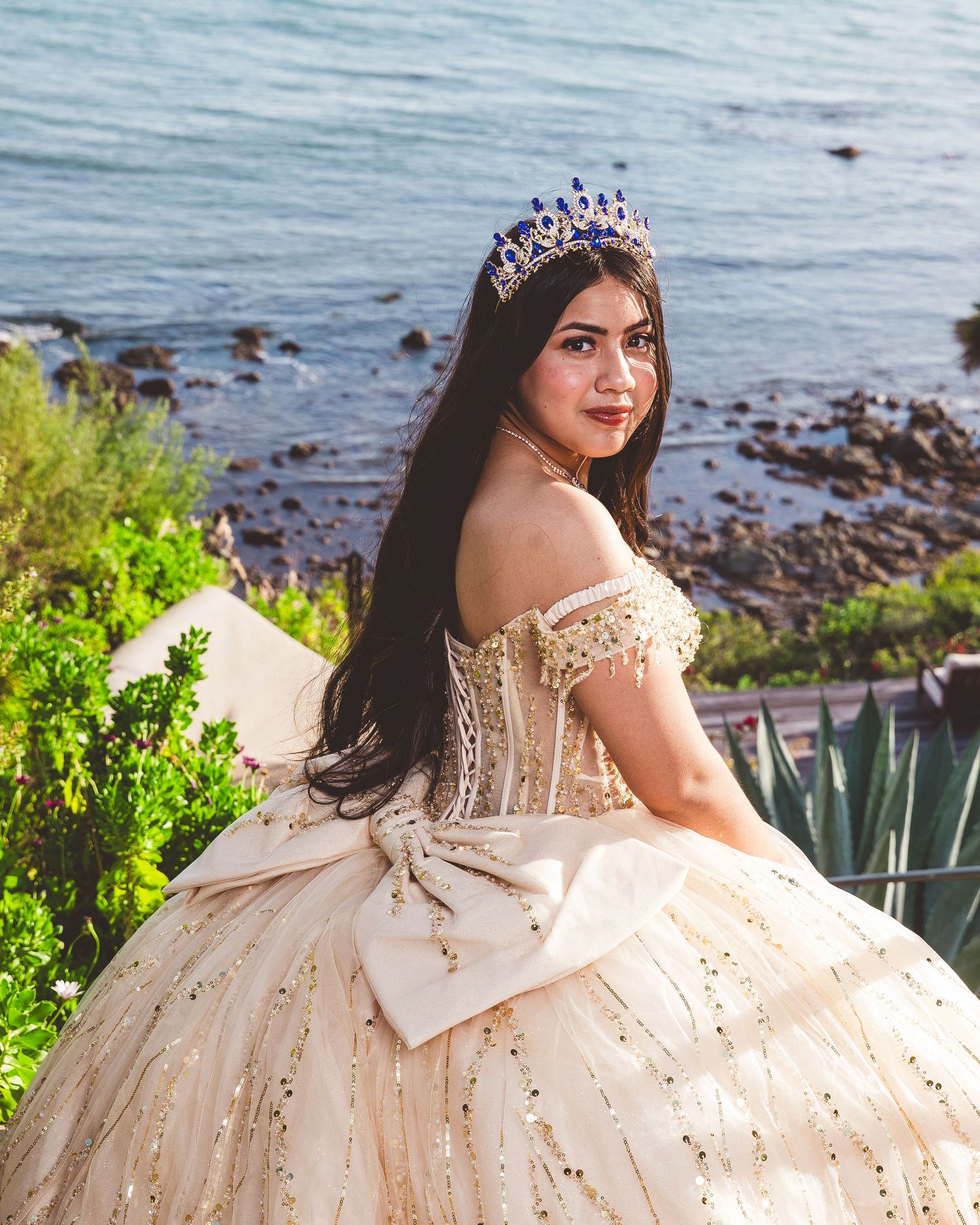 A sparkling champagne-colored quinceañera ball gown with off-shoulder details and crystal tiara photographed by the ocean.