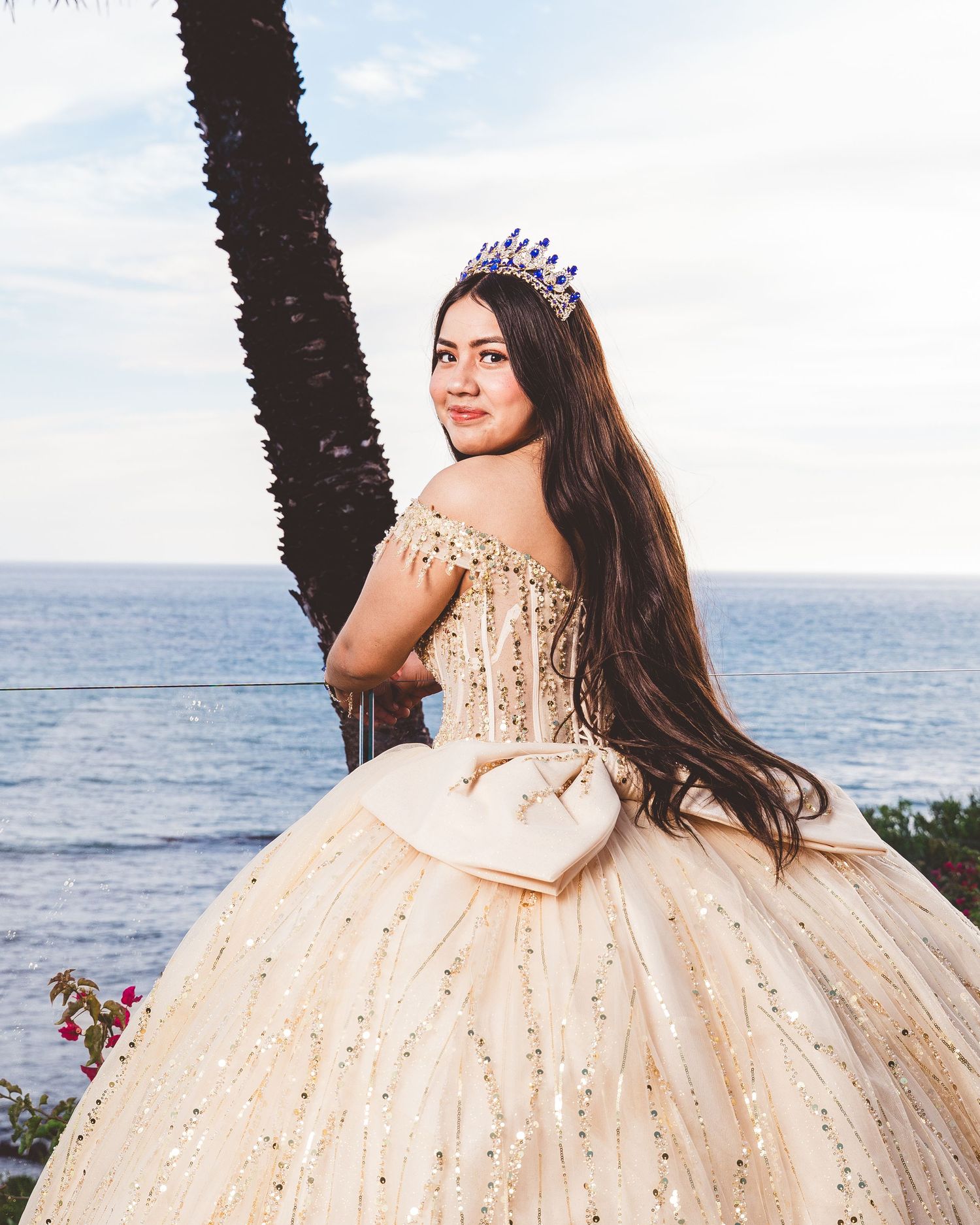 A flowing champagne-colored ballgown with sparkly details is worn at a scenic oceanside location with a palm tree.