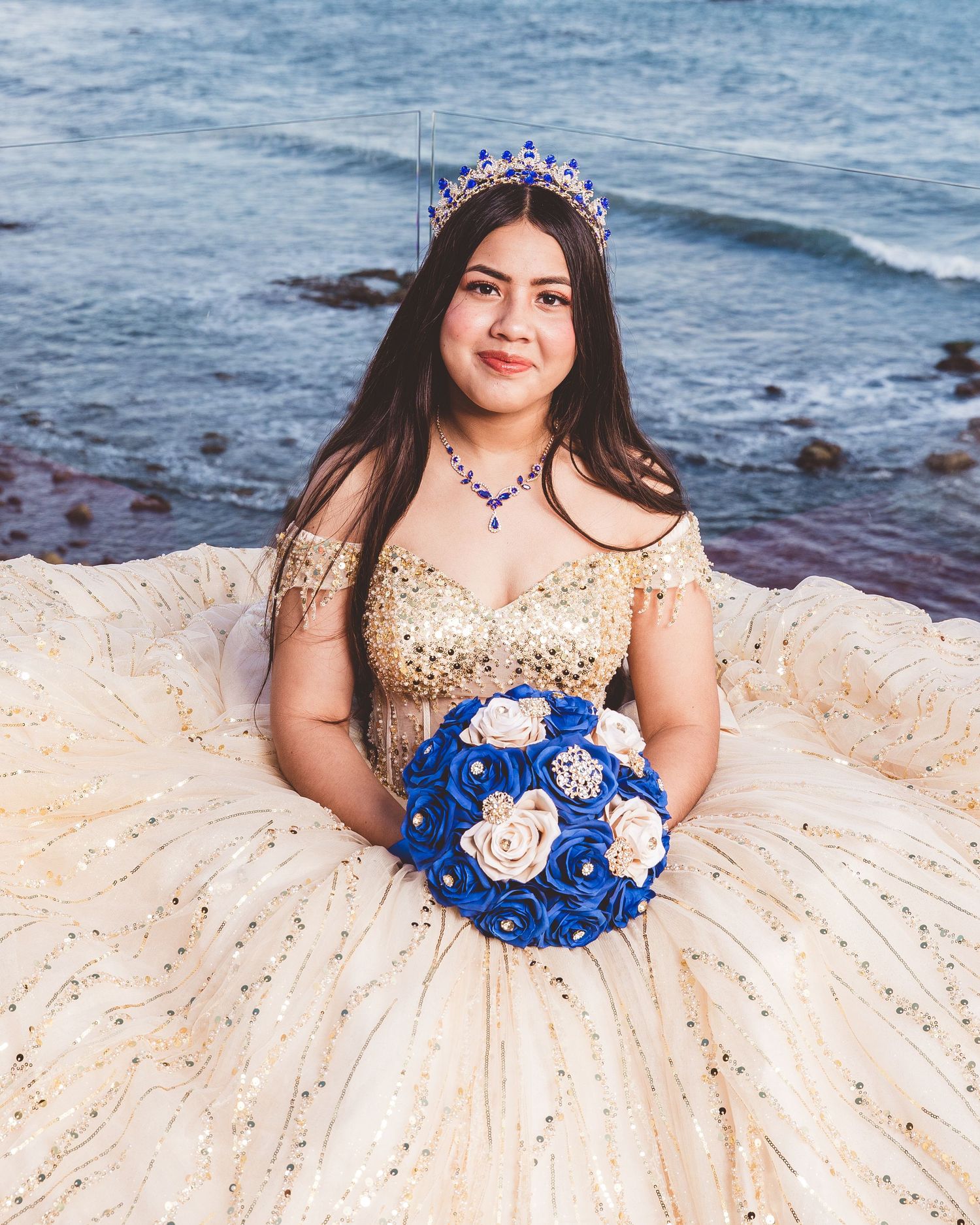 A person in a champagne-colored quinceañera dress with blue floral bouquet poses on a rocky beach at sunset.