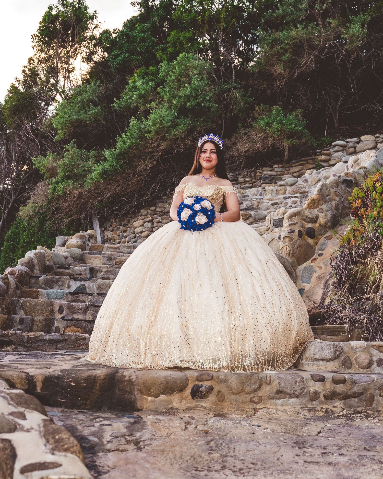 A person in a sparkly cream ballgown poses on stone steps surrounded by lush greenery at sunset.