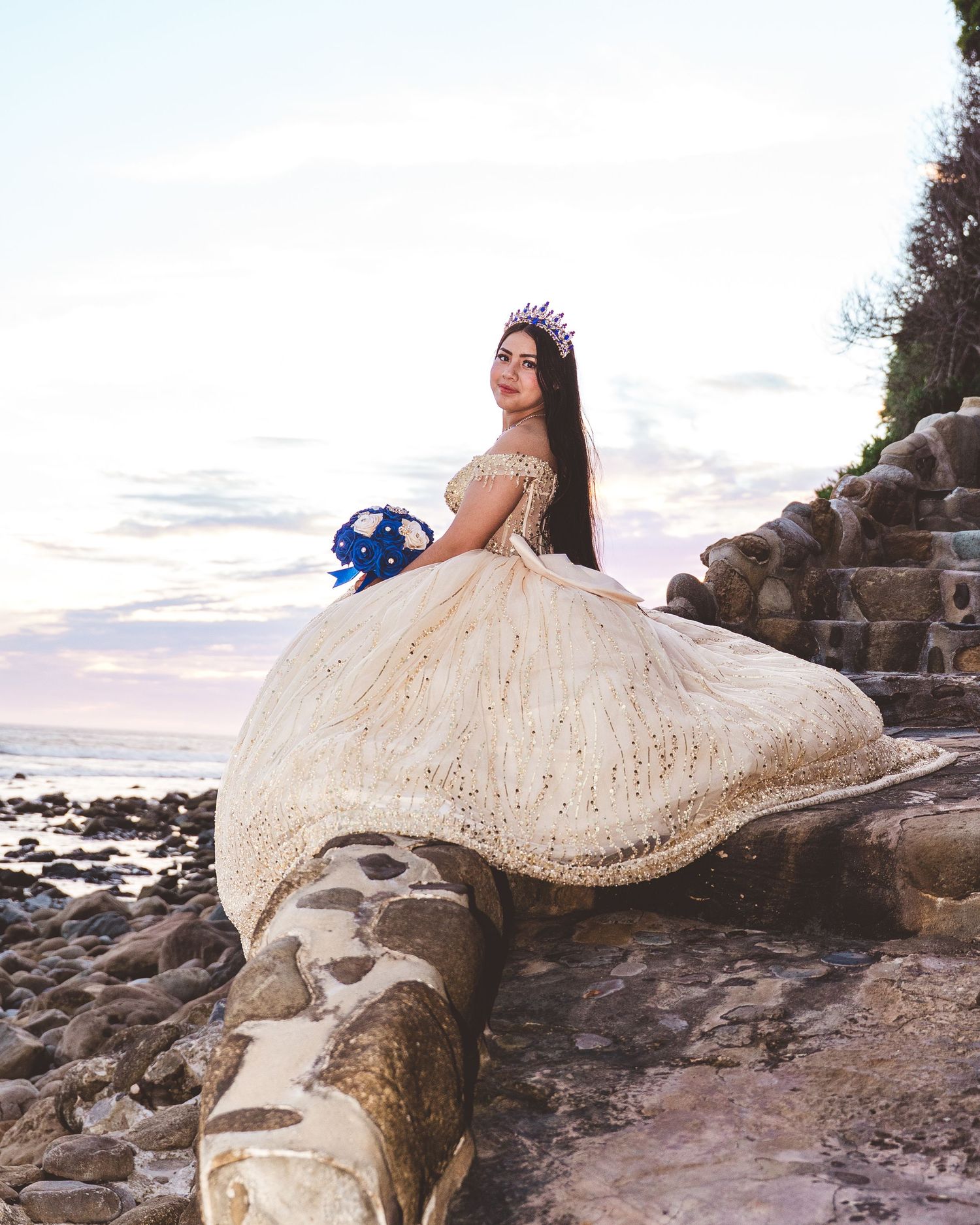 A bride in a flowing cream lace dress poses gracefully on rocky coastal terrain at sunset.