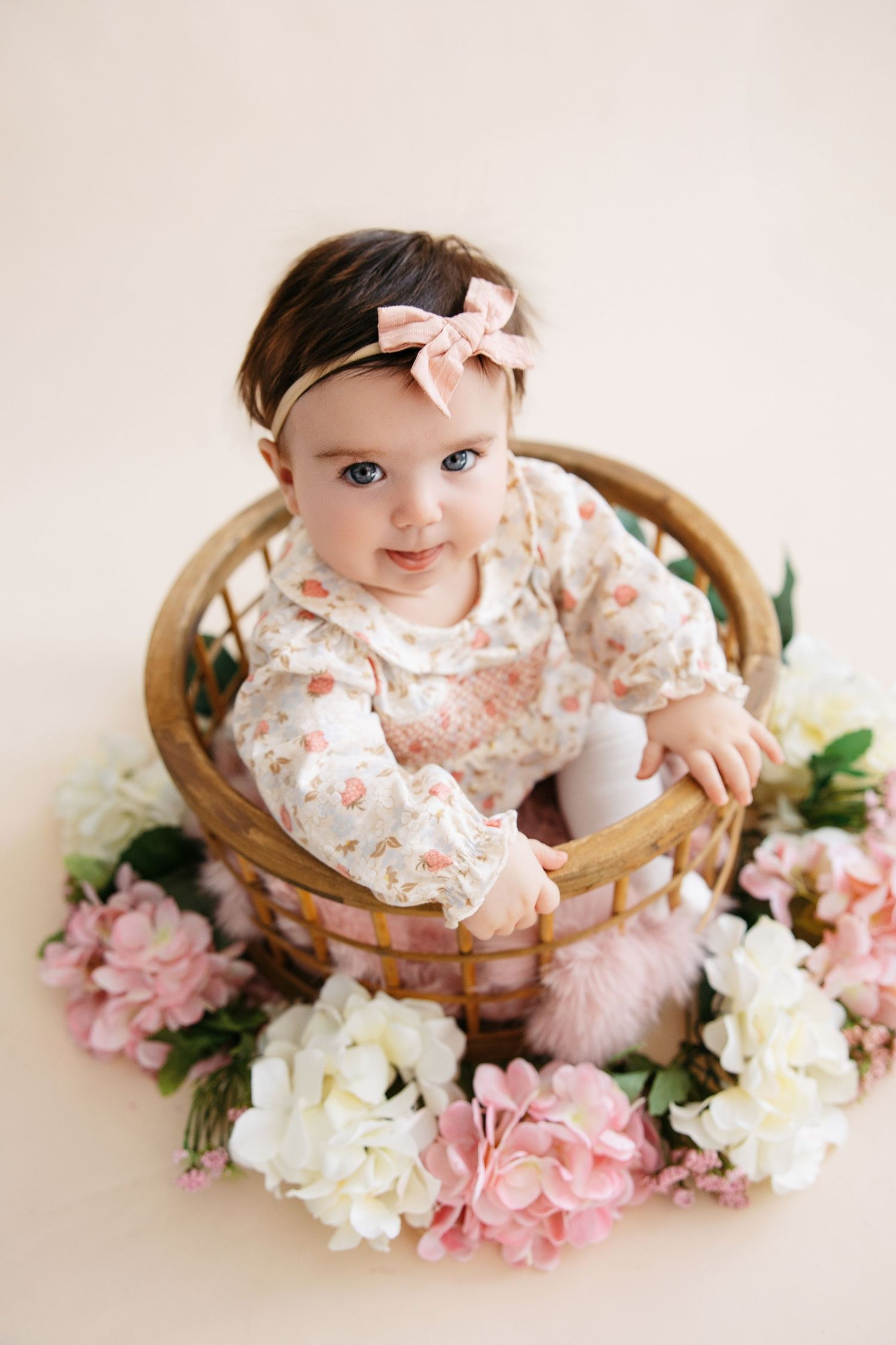 A baby in a floral dress sits in a wooden bowl surrounded by pink and white flowers against a cream background.