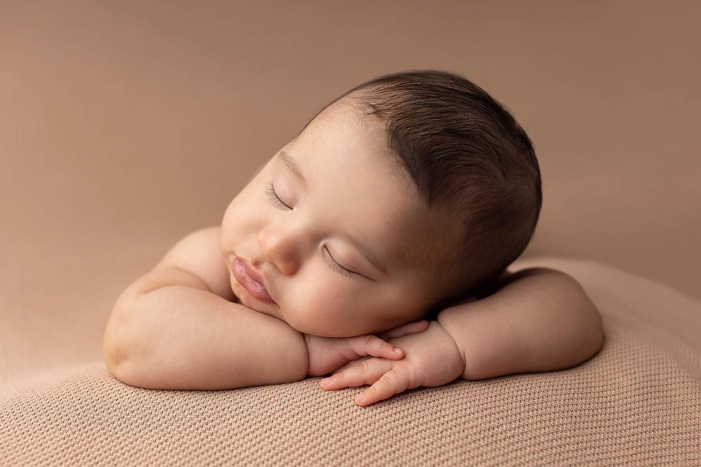 Peaceful sleeping newborn baby resting with folded arms on soft beige textured backdrop in studio lighting.