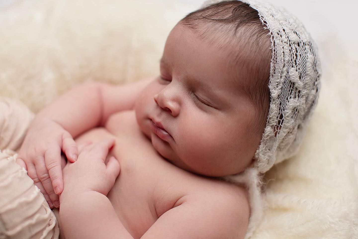 Peaceful sleeping newborn baby wearing delicate white lace bonnet resting on cream colored blanket.