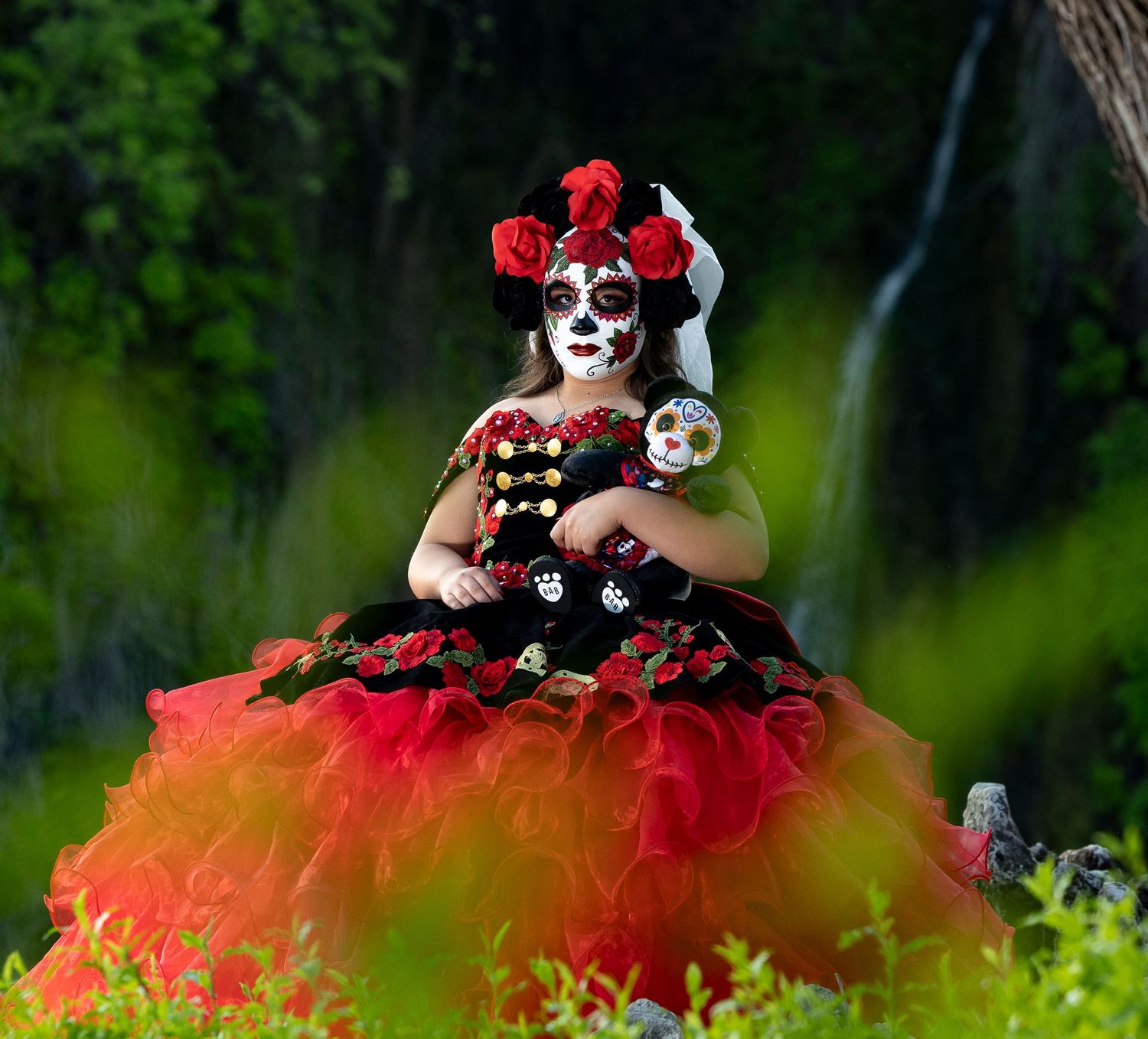 A figure in Day of the Dead makeup and red floral crown poses in a dramatic red and black tulle dress outdoors.