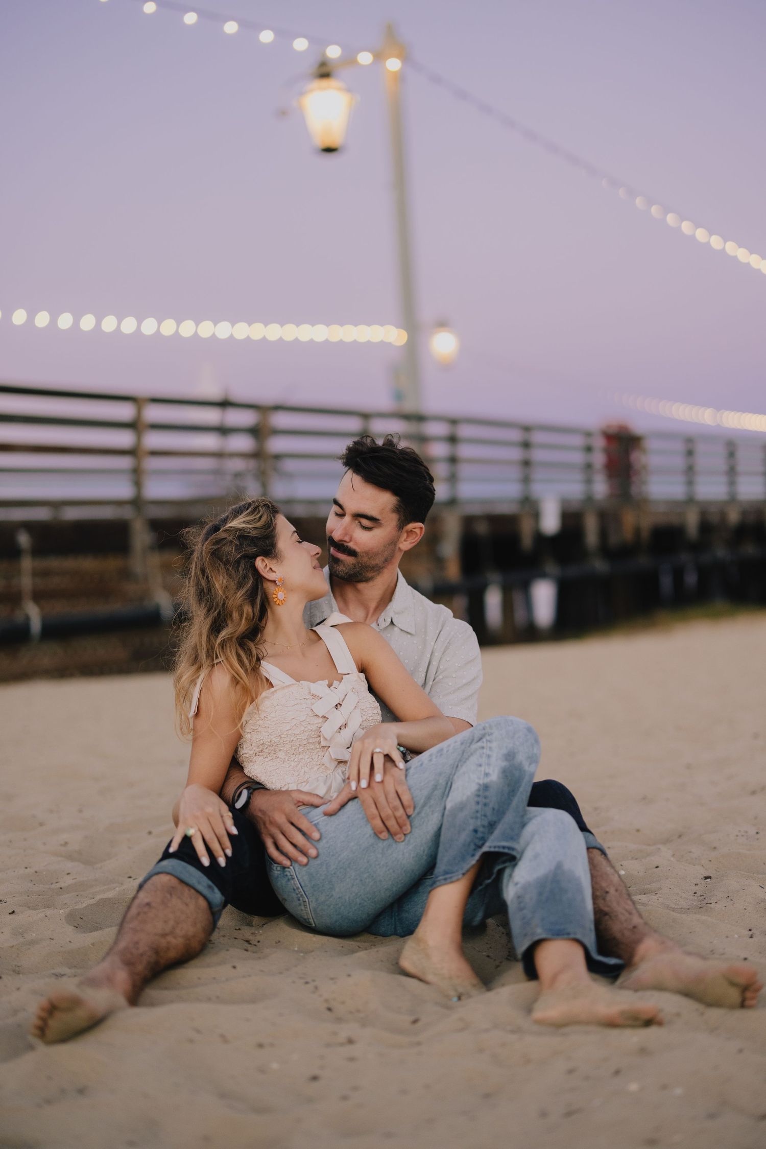 Couple sits closely together on sandy beach at dusk with string lights glowing behind them.