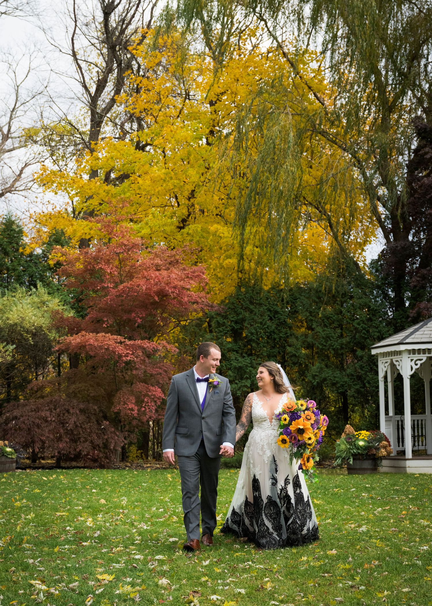 Wedding couple walks through vibrant autumn foliage with yellow and red maple trees in outdoor garden setting.