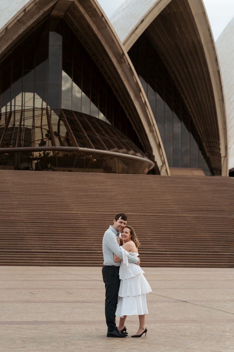 A couple hugs in front of the steps of the Sydney Opera House in a series of romantic poses during their engagement photoshoot in sydney