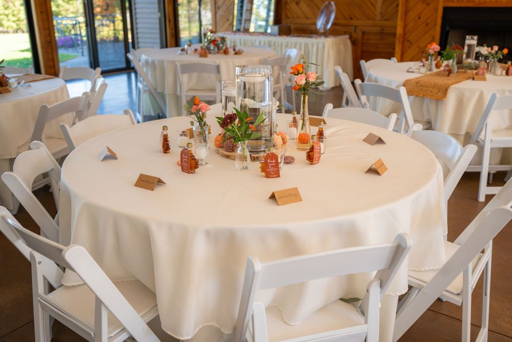 Wedding reception table setup with white tablecloth, wooden chairs, and centerpiece decorations in rustic venue.