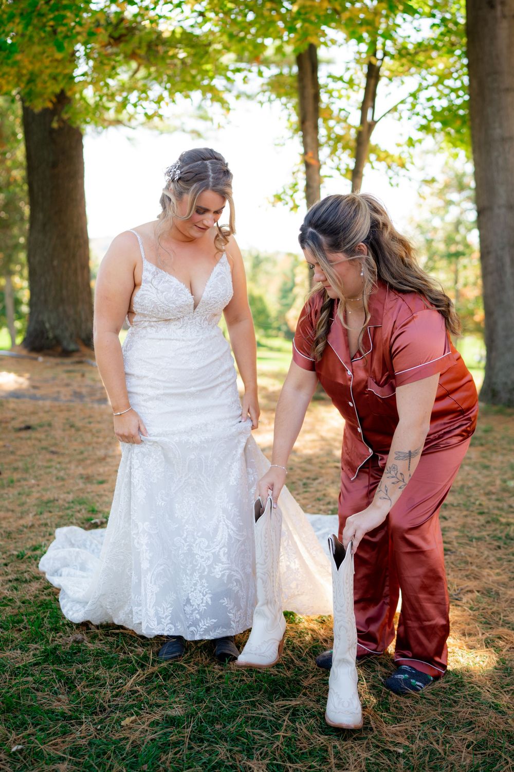 A couple in wedding attire helps adjust a white boot in a wooded outdoor setting during a photo session.