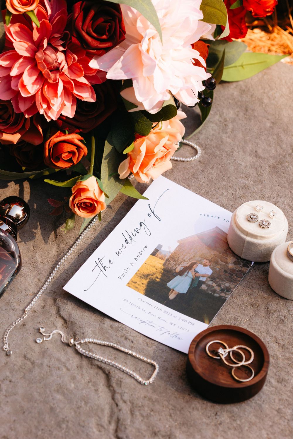 Wedding details flatlay with colorful flowers, rings in wooden dish, invitation card and decorative elements on grey surface.