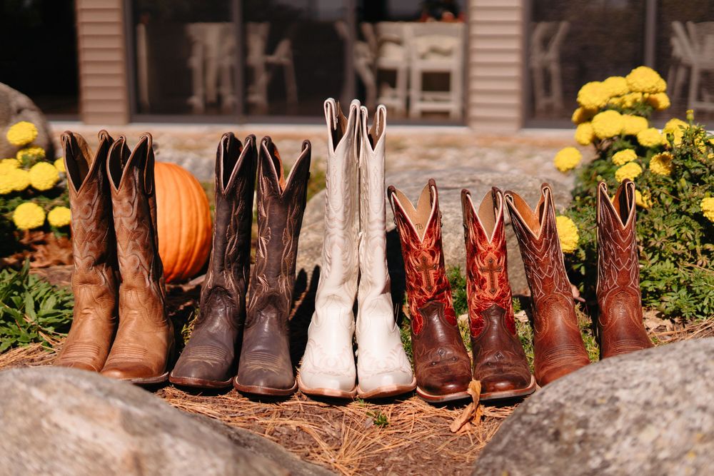 A row of cowboy boots in various colors and styles lined up outdoors with a pumpkin and yellow flowers in the background.