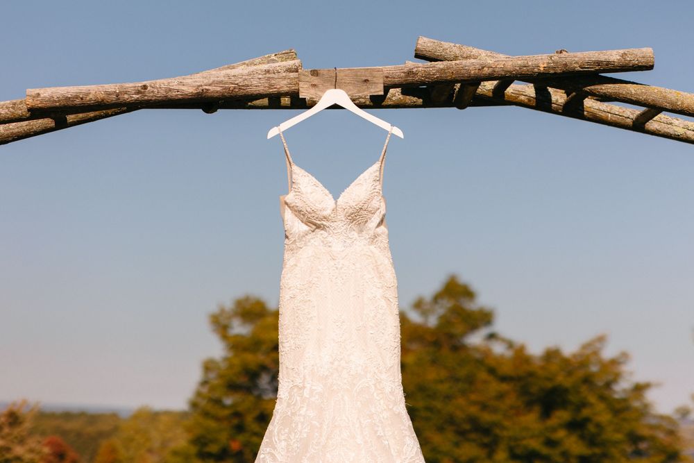 A white lace wedding dress hangs from a rustic wooden branch against a blue sky backdrop.