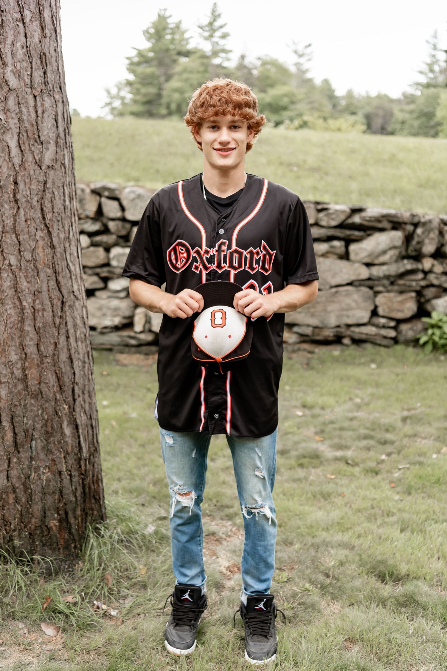 A person wearing a black baseball jersey and ripped jeans stands outdoors near a stone wall and pine trees.