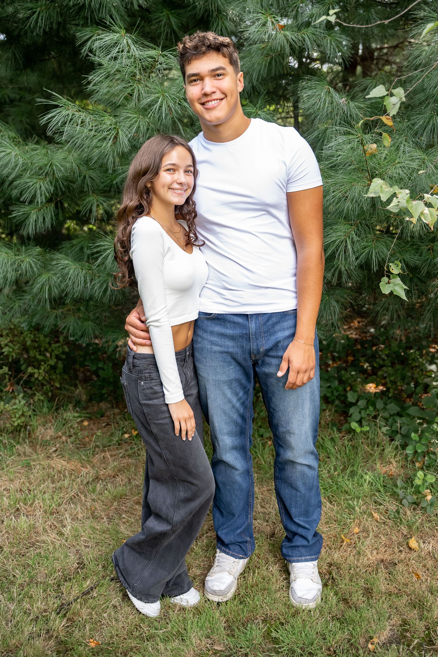 A couple stands together outdoors wearing casual white shirts and jeans against a backdrop of green foliage.