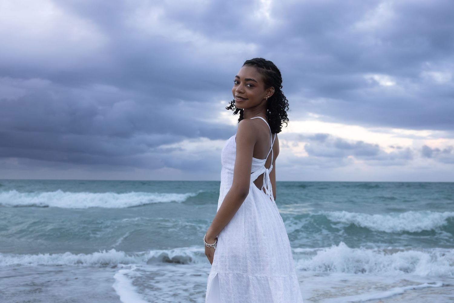 Young woman in white backless dress posing on beach with dramatic cloudy ocean backdrop.