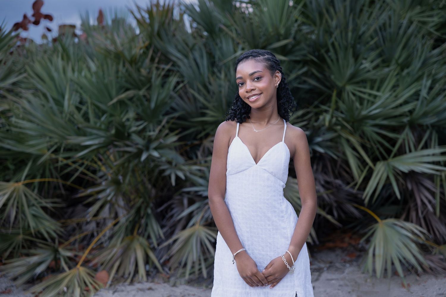Young woman in a white dress smiling in front of lush tropical green palm plants outdoors.