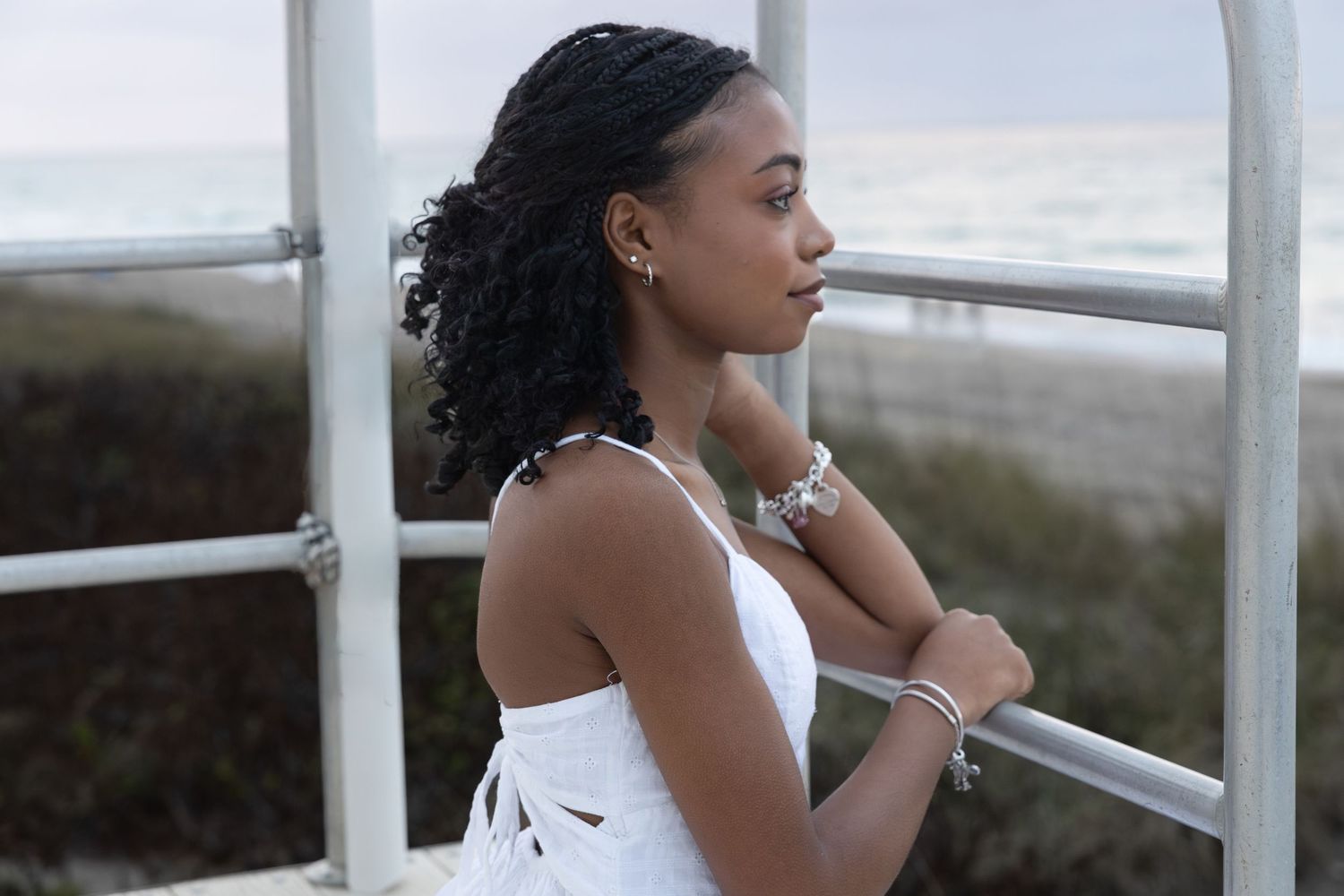 Elegant young woman in white dress leaning on a railing with ocean view in background.