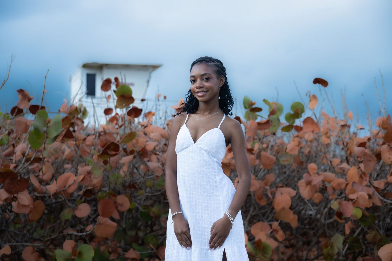 Young woman in white dress standing among autumn coastal shrubs under cloudy sky.