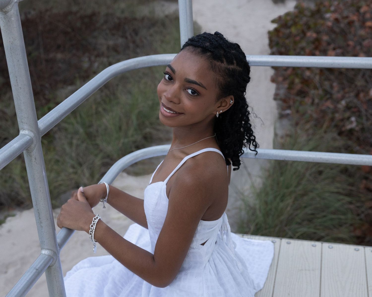 Smiling young woman in white sundress sitting on beach boardwalk stairs.