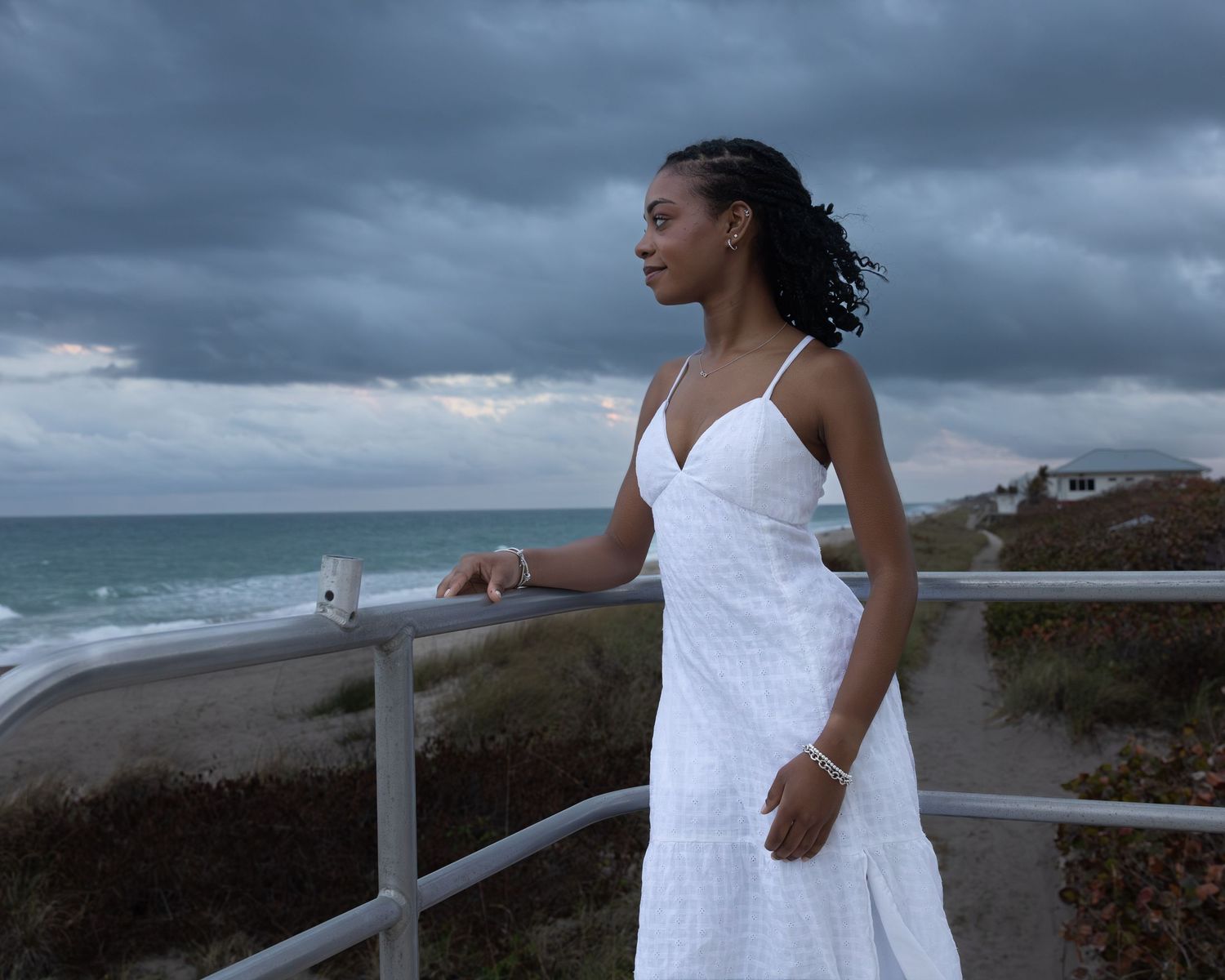 Young woman in white sundress poses gracefully on beach boardwalk railing with stormy ocean backdrop.