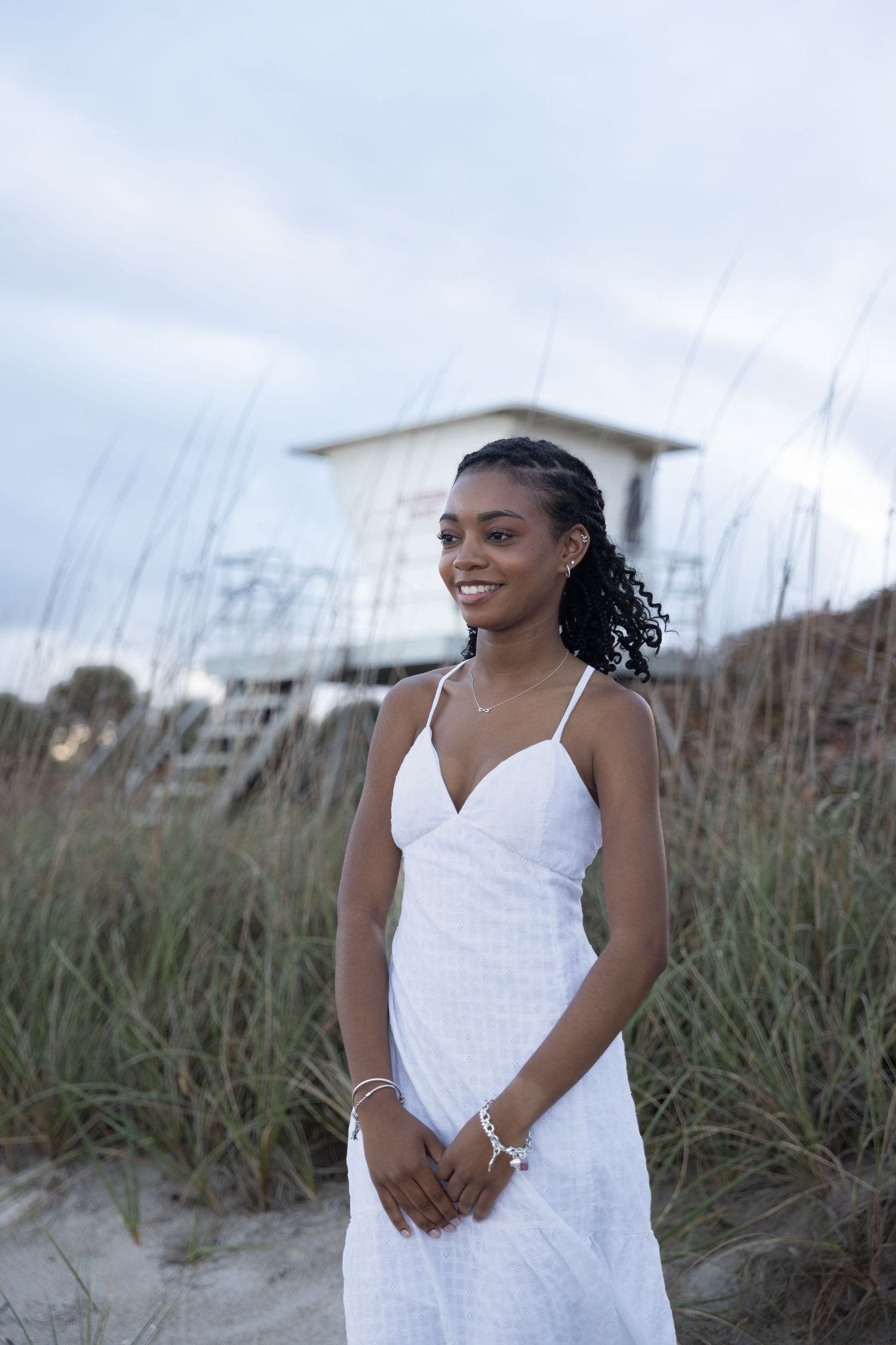 Smiling young woman in white sundress stands on sandy beach near lifeguard tower and sea oats.