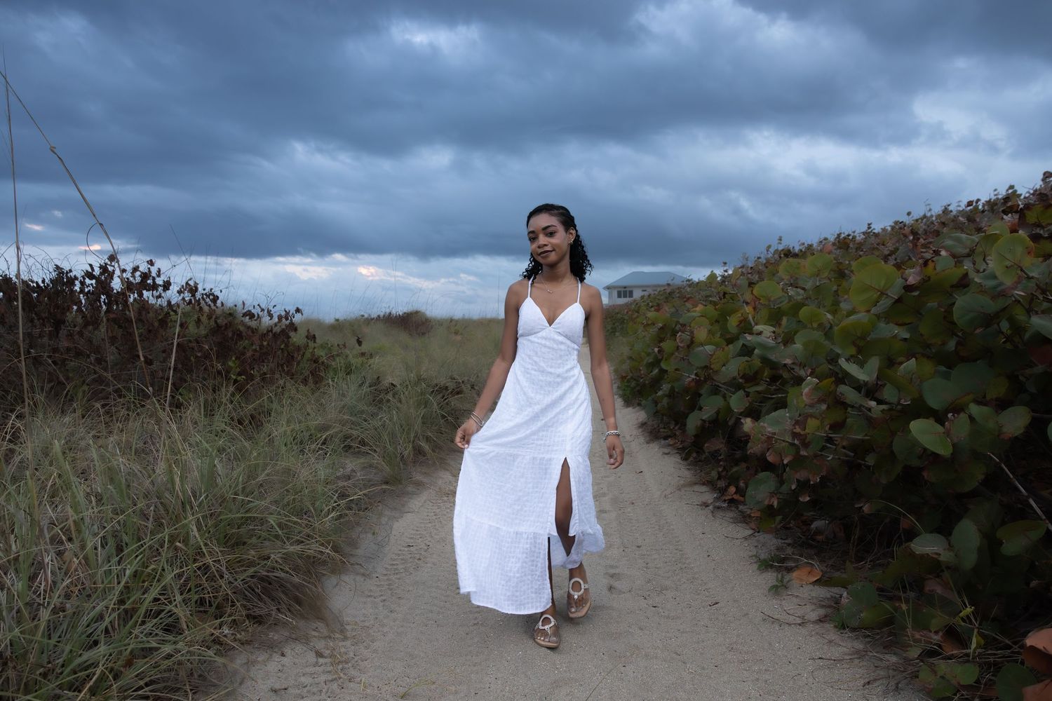 Young woman in a white maxi dress walking a sandy beach path under dramatic cloudy skies.