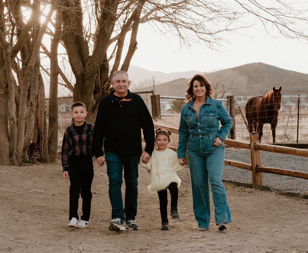 A family walks together on a dirt path with horses and bare trees visible in the rural winter landscape.
