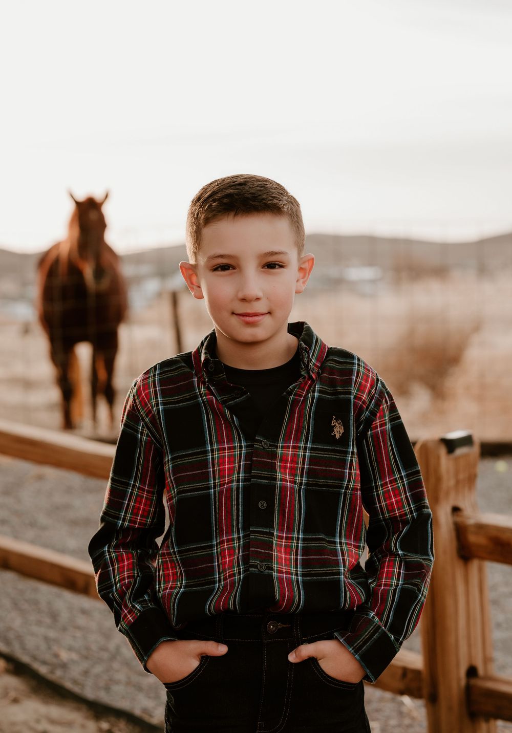 A child in a plaid shirt poses outdoors with horses visible in the background at sunset.