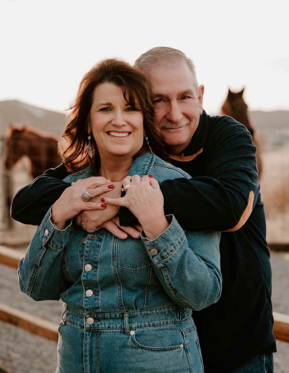 A couple embraces in a romantic outdoor setting with horses visible in the background at sunset.