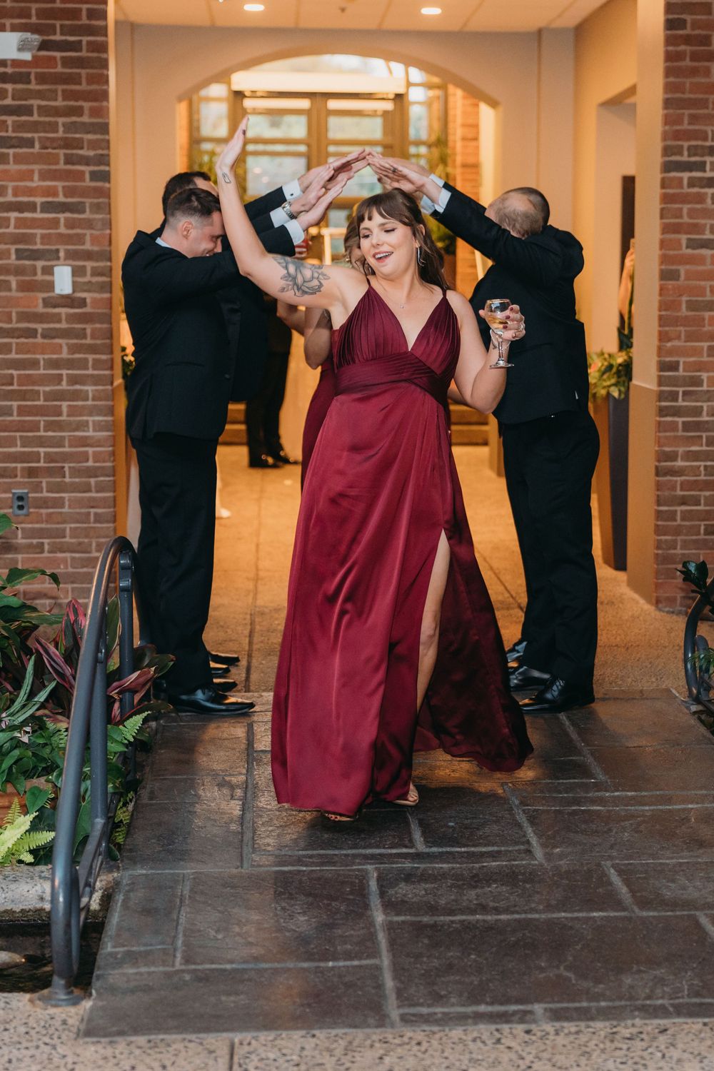 Wedding party members in burgundy dresses and black suits dance together in a brick-lined hallway.