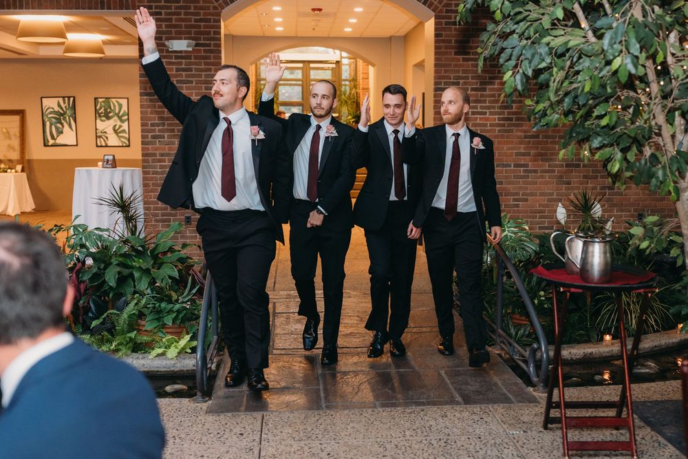 A wedding party in formal black attire walks together outside a brick building with warm lighting.