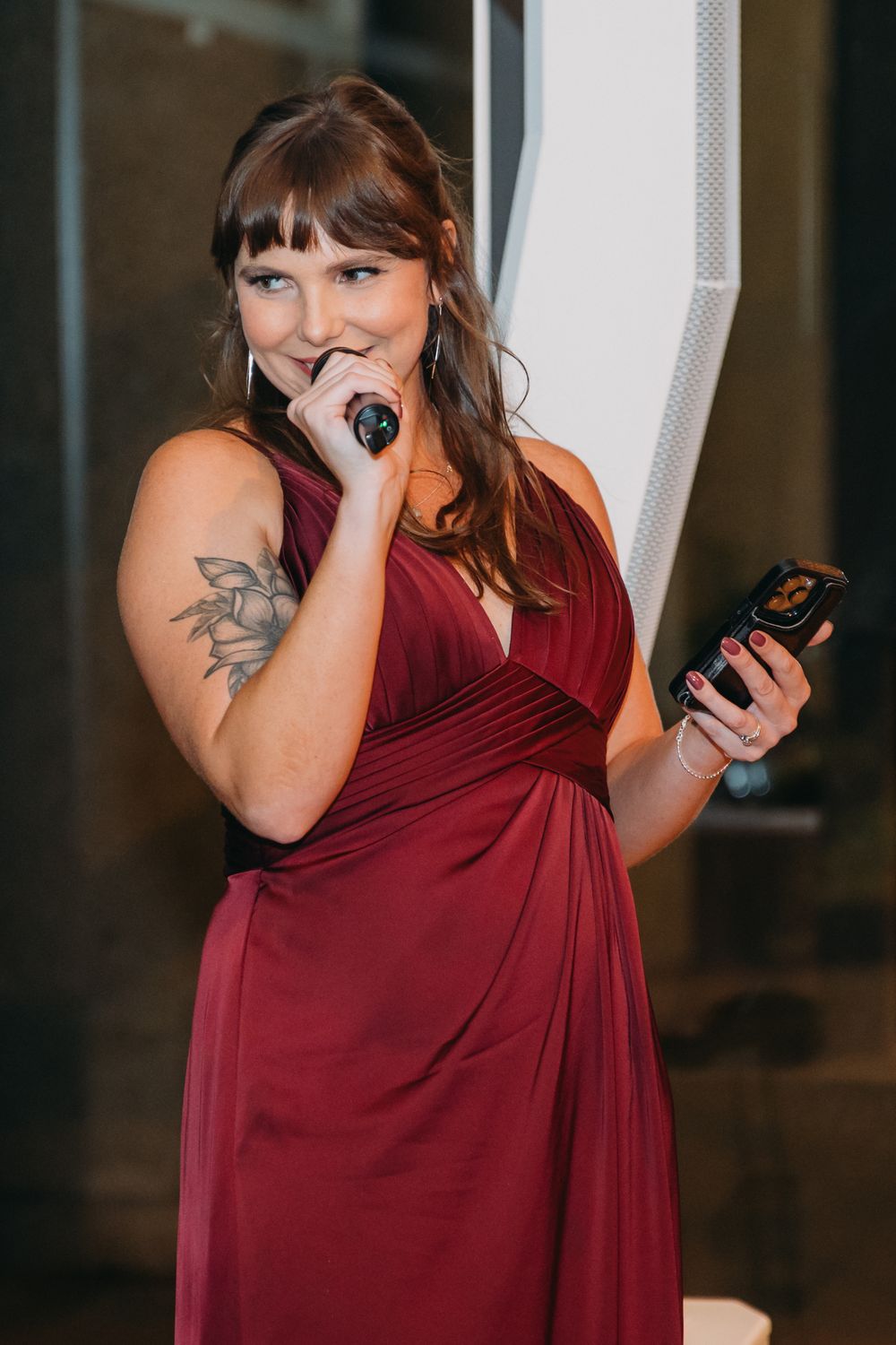 Person in burgundy dress holding a phone and covering their mouth in an emotional moment during a wedding reception.