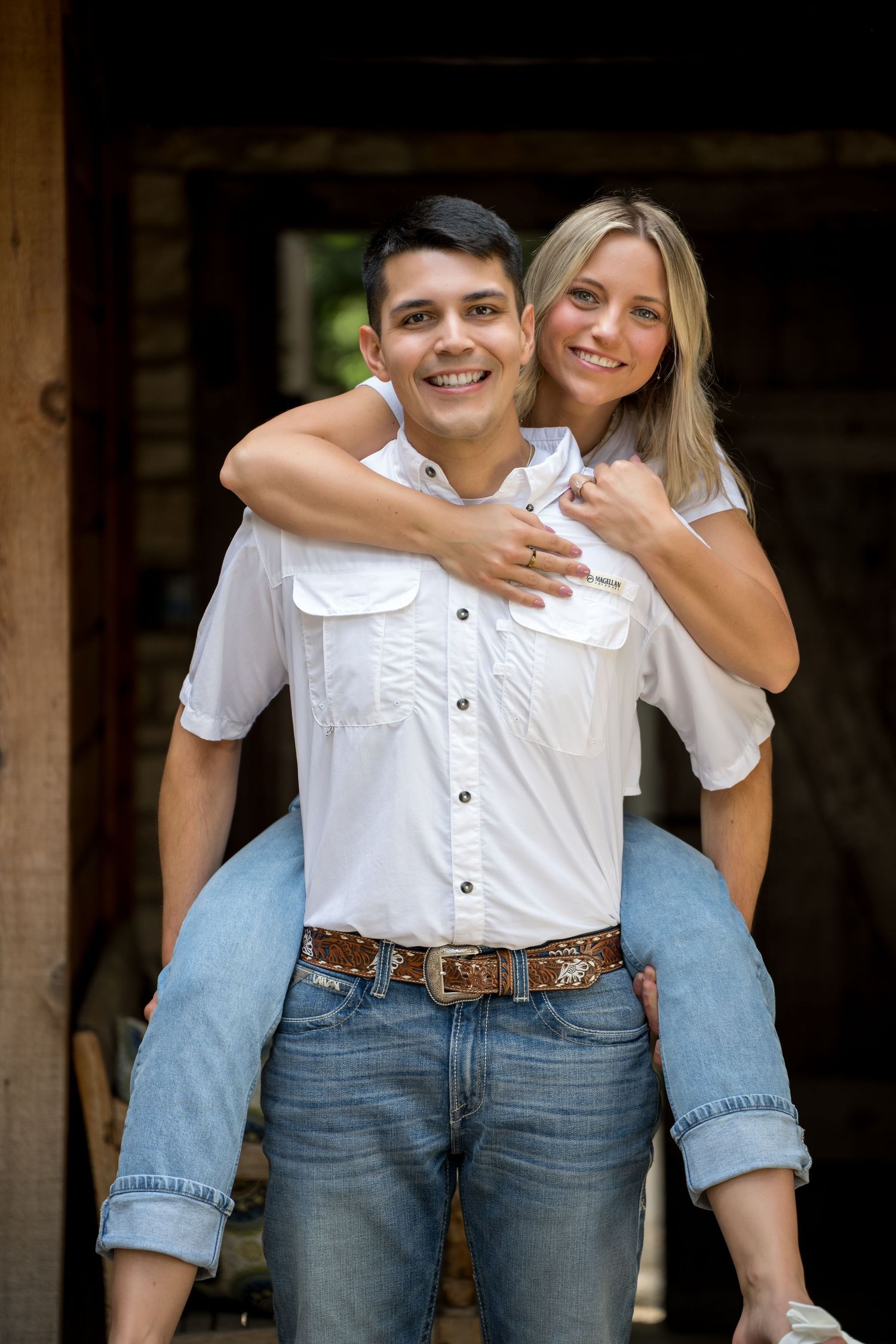 A smiling young couple in white shirts and jeans, with the man giving the woman a piggyback ride outside a barn.