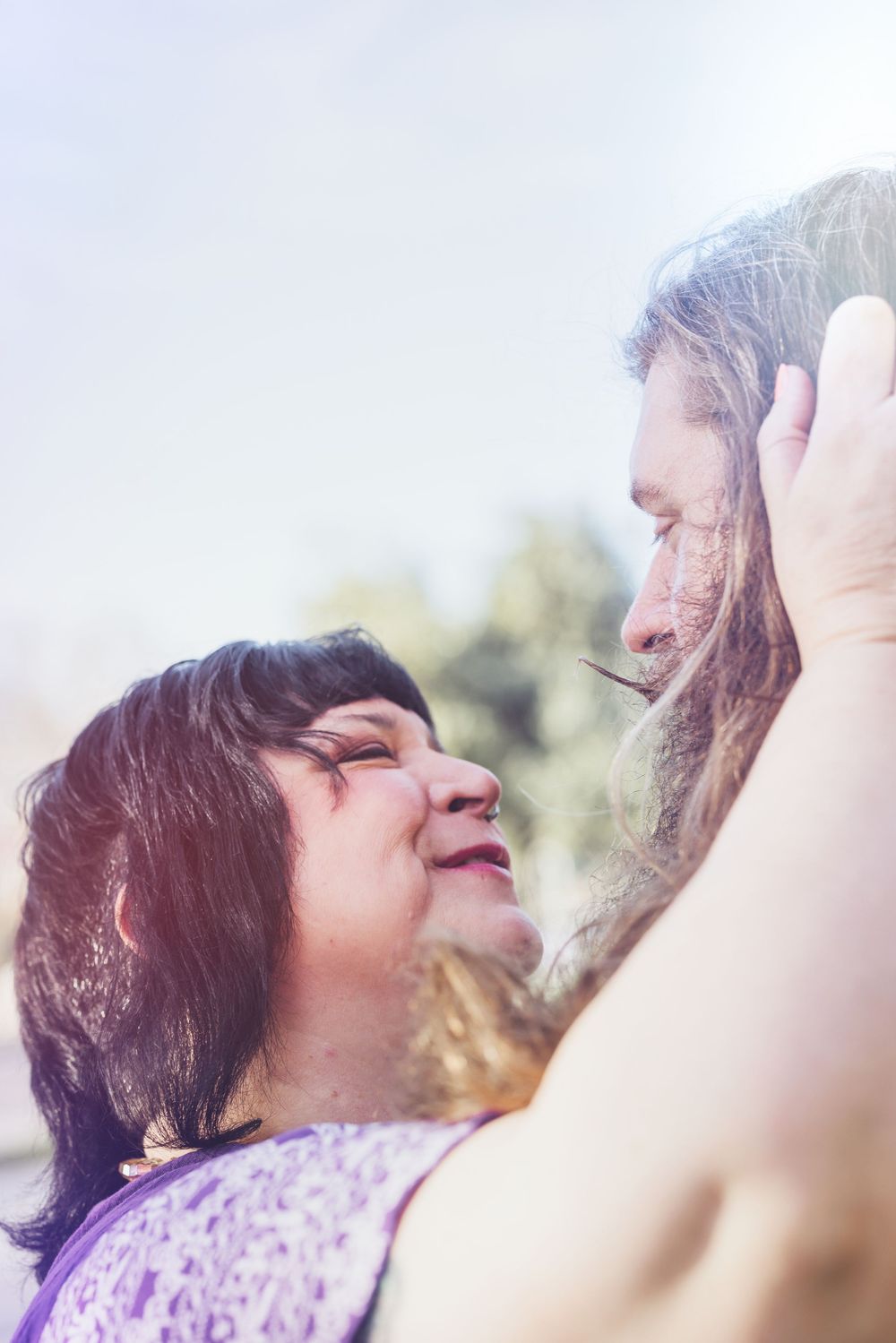 Woman in purple lace top laughs joyfully with arms raised against a sunlit outdoor background.