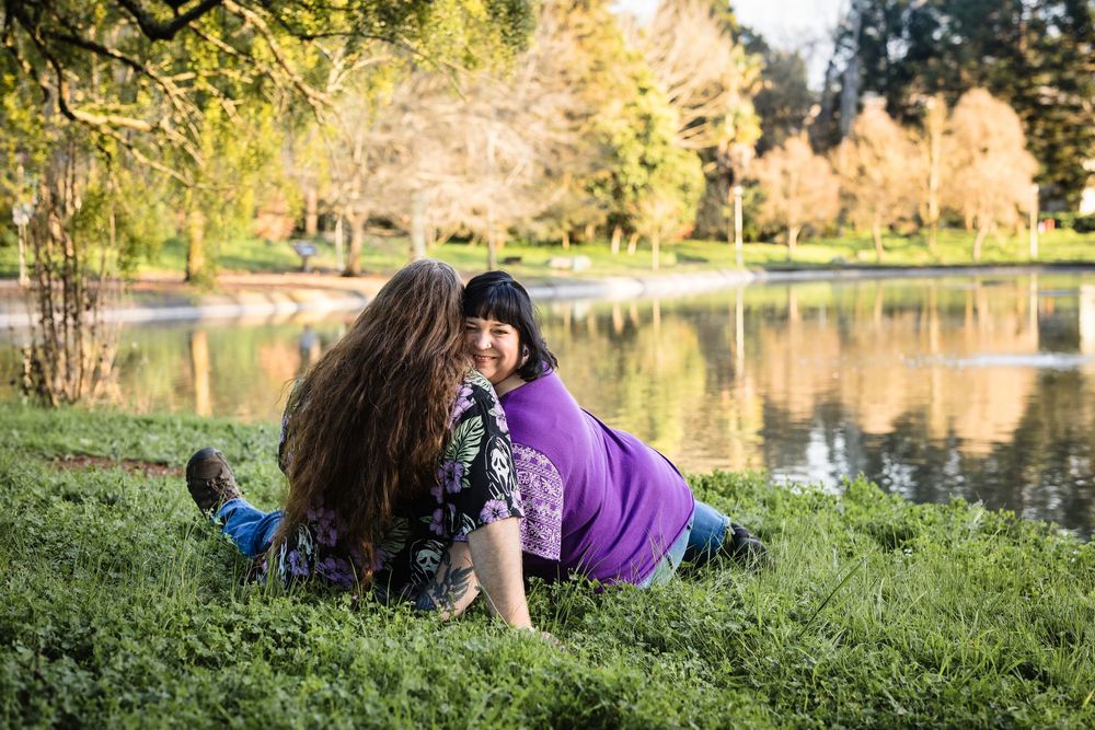 A couple relaxes on grassy lakeside hill during golden hour with autumn trees and water in background.