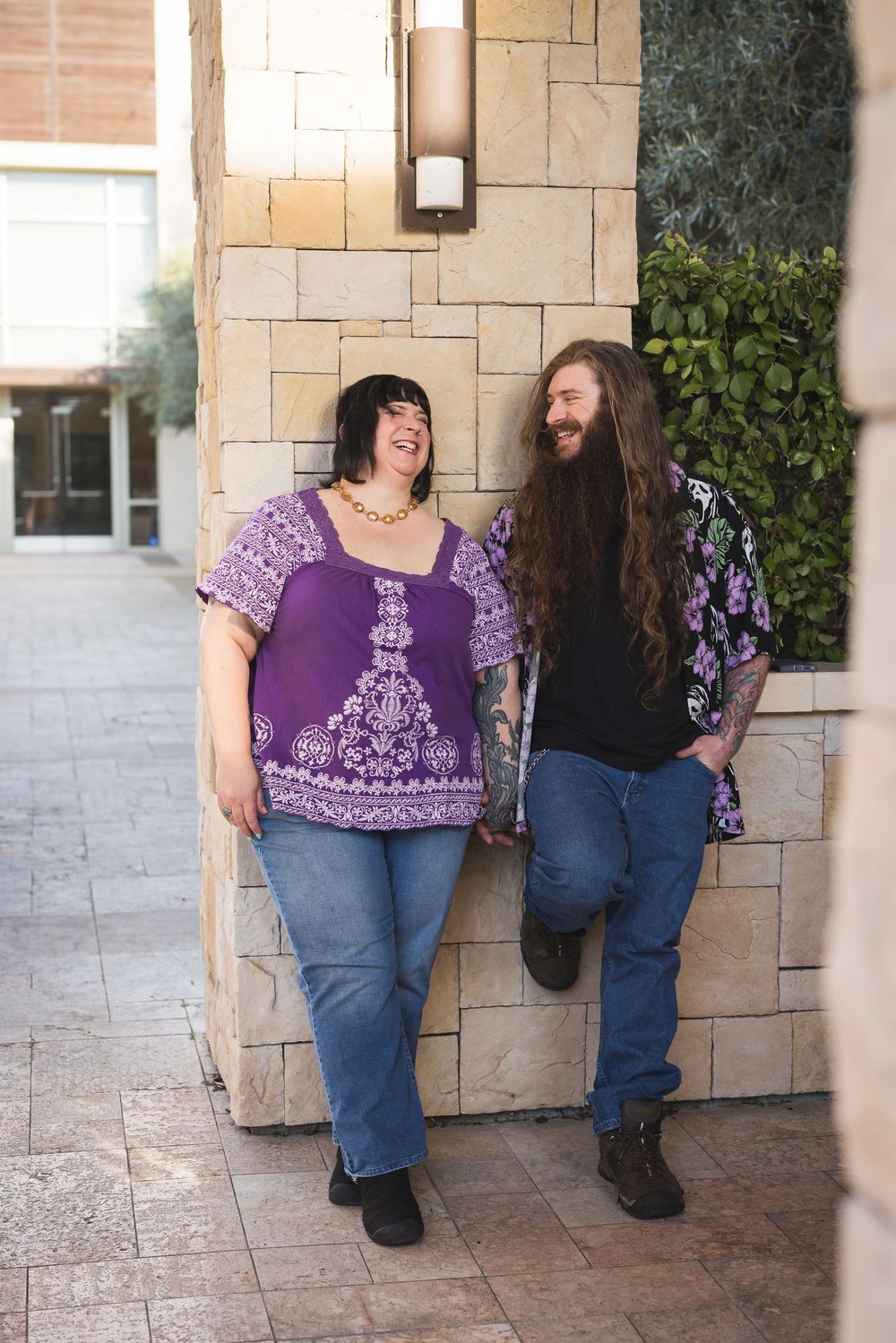 Two people share a laugh while leaning against a stone wall, wearing casual clothing and enjoying a candid moment.